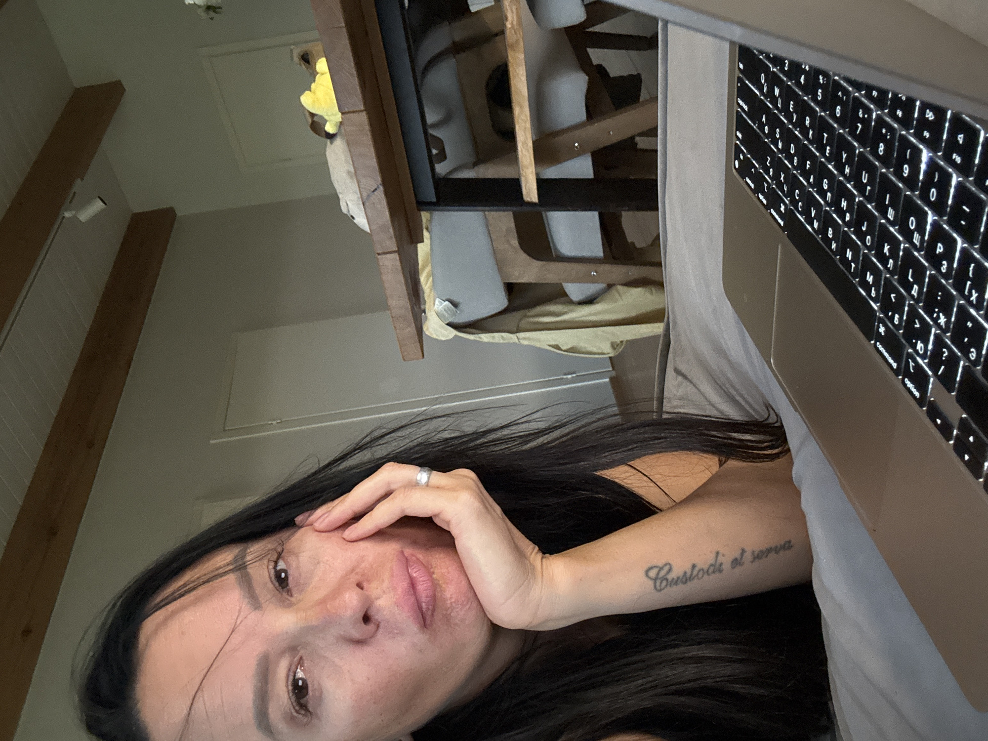 A woman with long, dark hair is lying on a bed, resting her head on her hand, and looking at the camera with a neutral expression. She is wearing a black tank top and has a tattoo on her left forearm. A silver ring is visible on her right hand. In the background, a wooden table with chairs and a yellow stuffed animal is partially visible. A laptop is placed in front of the woman, its keyboard illuminated. The scene appears to be indoors, possibly a bedroom or living area, with wooden beams and a white ceiling. The image does not provide any specific details or context to determine its location in Chaikovsky, Russia.