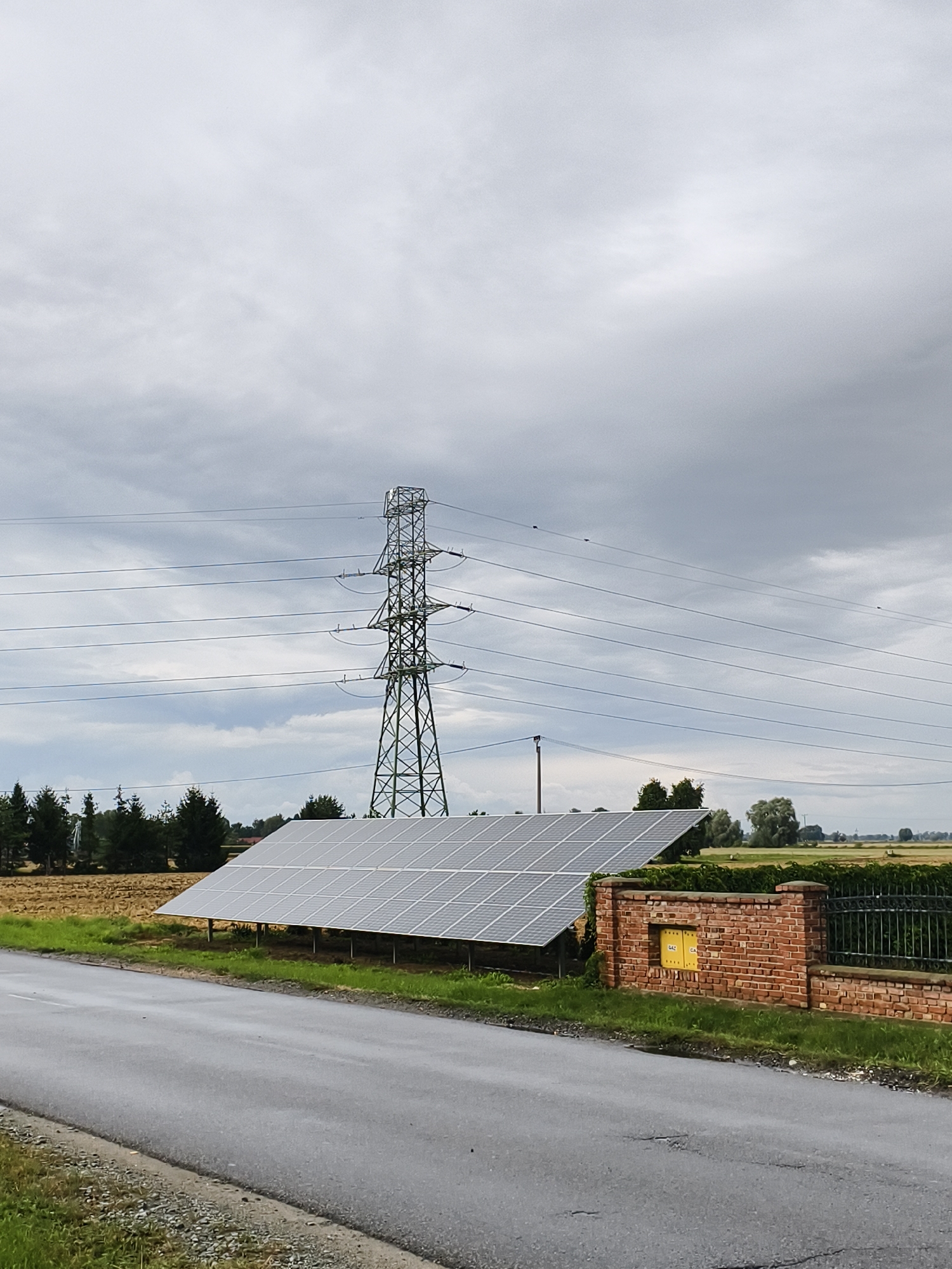The image depicts a scene in Tczewskie Łąki, Poland, featuring a large solar panel array situated in a field. A tall electricity pylon with multiple power lines stretches across the frame, dominating the background. To the right of the solar panels, a brick wall with a yellow utility box is visible, bordering a metal fence. The environment includes a paved road in the foreground and vegetation surrounding the solar installation. The sky is overcast with clouds. There are no people or animals visible in the image.