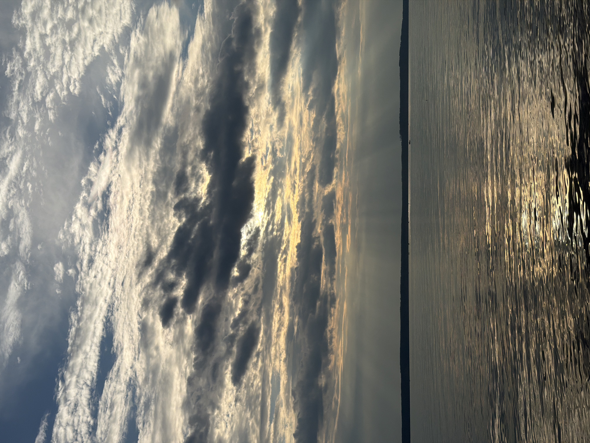 The image depicts a wide shot of a body of water under a cloudy sky. The water is calm with a slight ripple, reflecting the light from the sky, creating streaks of yellow and white on its surface. A dark, distant shoreline with trees is visible on the horizon. The sky is filled with a variety of clouds, ranging from thin, wispy white clouds to darker, more ominous gray ones. Rays of sunlight pierce through some of the clouds, creating dramatic shafts of light that extend towards the water. The overall impression is one of a serene, yet dramatic natural landscape. The location is identified as Stepanovo, Russia.