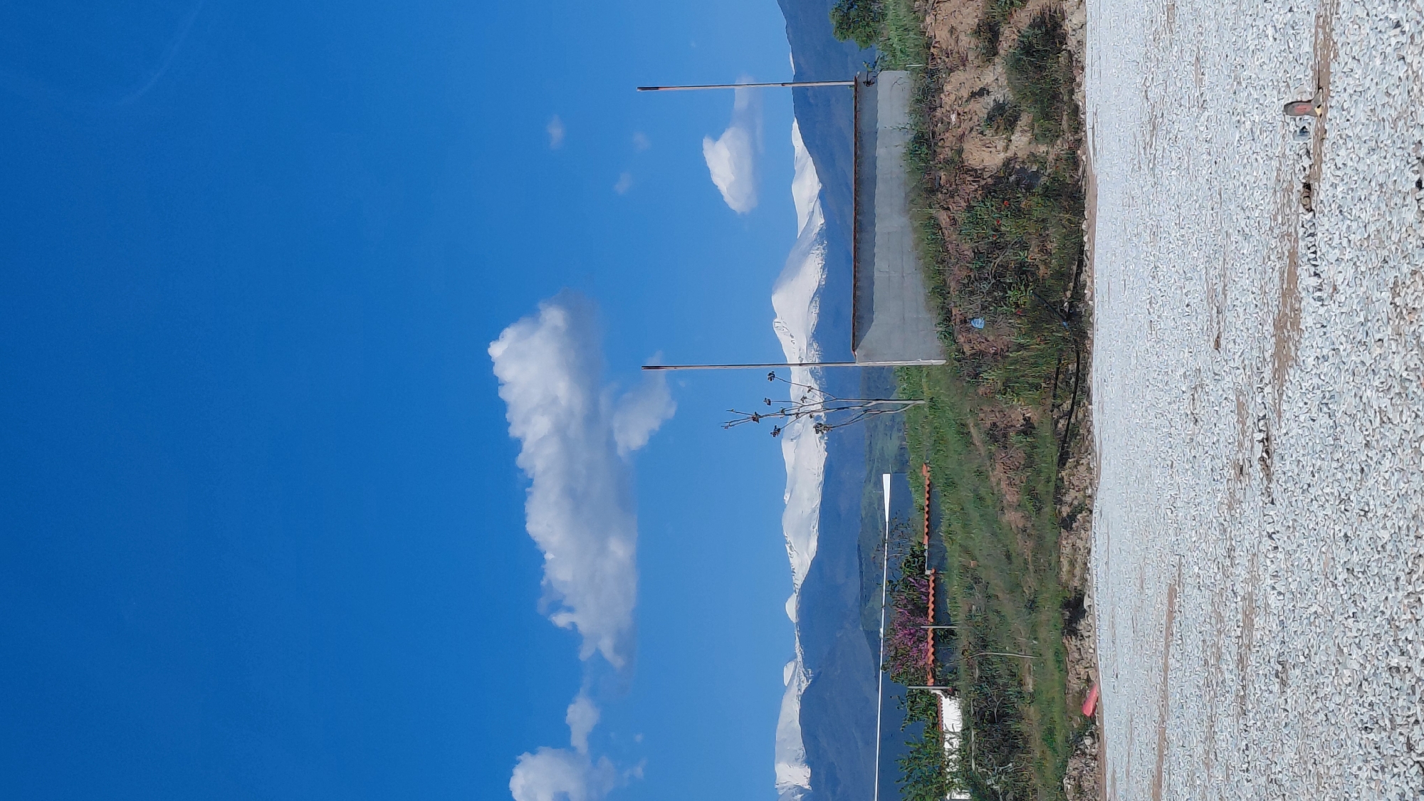 The image shows a landscape featuring a clear blue sky with fluffy white clouds on the left. In the center, there are snow-capped mountains in the background. In the foreground, there's greenery and some foliage, with a building featuring a red-tiled roof partially visible. On the right, the ground appears to be covered with gravel. Additionally, there are two vertical poles extending upwards, seemingly unrelated to the landscape. The orientation of the image is rotated 90 degrees counterclockwise. Location: Unknown, Unknown.