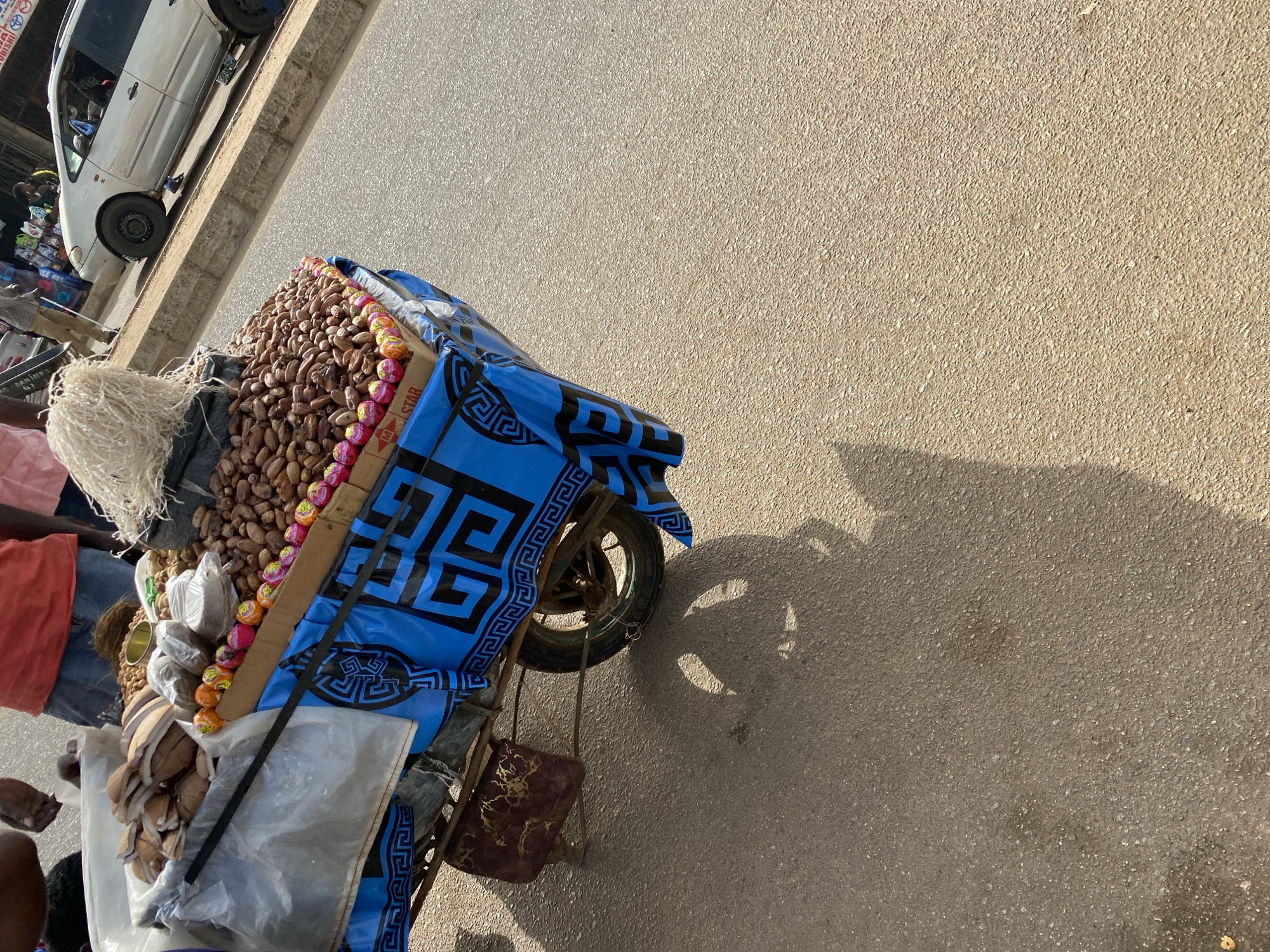This image, captured in Bukuru, Nigeria, depicts a street vending scene under bright sunlight, likely in the morning or late afternoon given the long shadows on the pavement. The primary focus is a vendor's tricycle or pushcart heavily loaded with merchandise.
The cart features a large pile of reddish-brown nuts, possibly kola nuts, prominently displayed. Along one edge of the nuts, rows of small, colorful, individually wrapped items, possibly candies or snacks, are arranged. The text "M STAR" is visible on their packaging. Other goods, including items in clear plastic bags (some resembling split coconuts), are stacked on the cart. A blue fabric with a black Greek key (Meander) pattern covers part of the cart, and a brown bag with gold patterns hangs from its side.
A person, presumably the vendor, is partially visible on the left, wearing an orange/red shirt and with what appears to be white hair or a head covering. In the background, a white car, possibly a minivan, is parked next to a raised curb, suggesting a roadside or market setting. Other blurry figures and market stalls are visible near the car, and a partially obscured sign in the distance reads "MITSUBISHI". The paved road, showing distinct shadows cast by the cart and other objects, occupies a significant portion of the frame. The image is taken from a tilted, slightly elevated perspective.