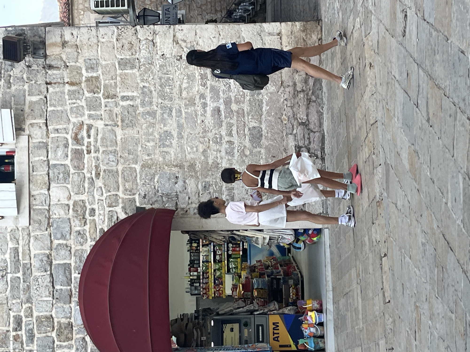 Three individuals walk on a cobblestone street in front of a stone building with a red awning covering a shop entrance. To the left, a shop interior is visible, stocked with shelves of various products, an ATM, and buckets of toys. The individual in the foreground on the left appears to be a young girl wearing a pink t-shirt and white shorts. She is followed by another young girl in a striped tank top and shorts, carrying white plastic bags. To the right, a woman in a dark blue shirt and shorts walks away from the camera, carrying a dark green bag over her shoulder. The stone building suggests an older architectural style. A sign for "JUNTA RESTAURANT" is visible on the right side of the image. The lighting indicates it is daytime.