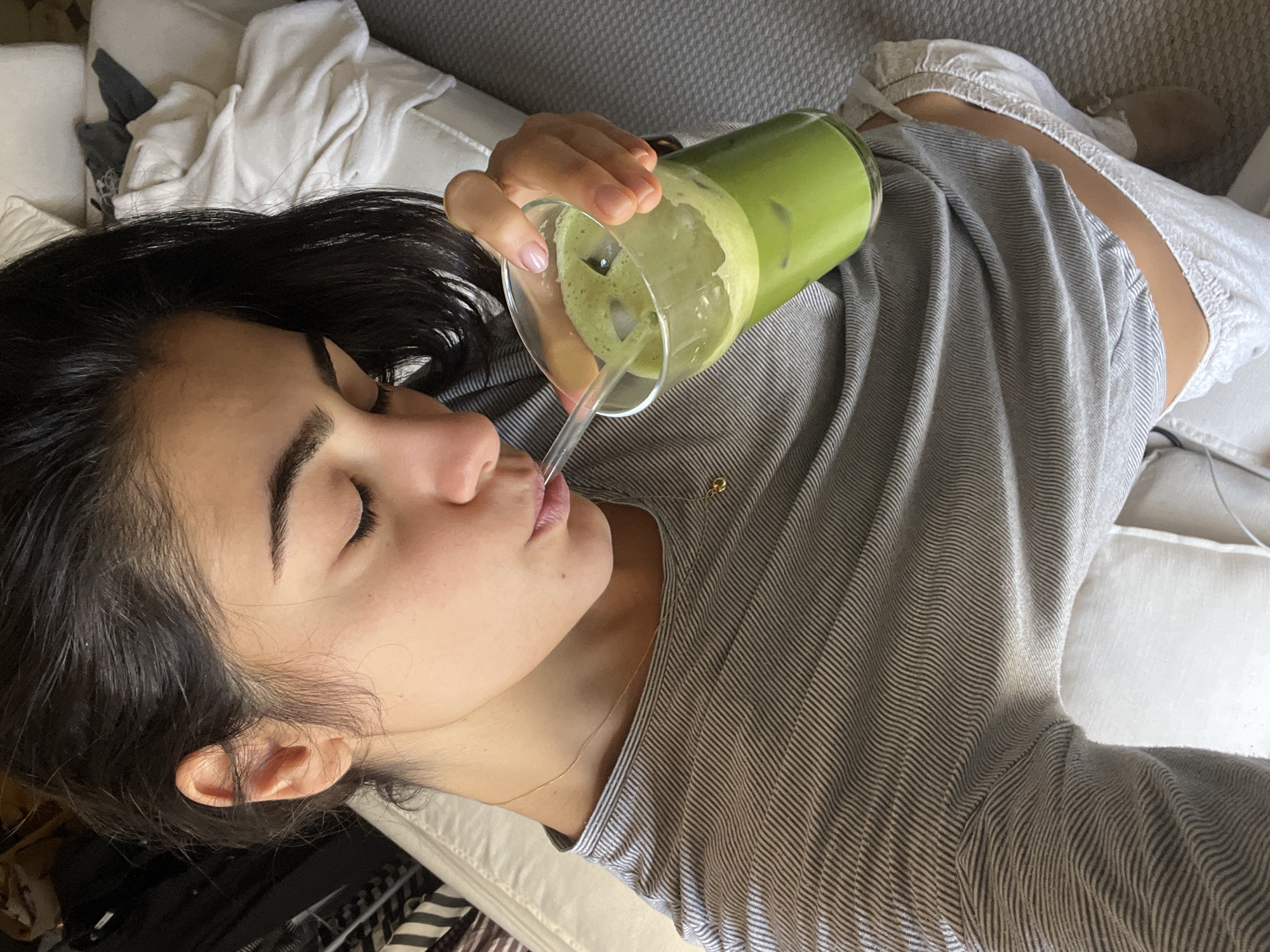 A woman with dark hair is lying on a light-colored couch, resting on her left side. She is drinking a green beverage, possibly matcha tea, from a tall glass filled with ice through a straw. She wears a gray striped shirt and a delicate gold necklace. Her eyes are closed. The scene appears to be indoors, likely in a home. There is no indication of the city of Paris, France, in the provided image.