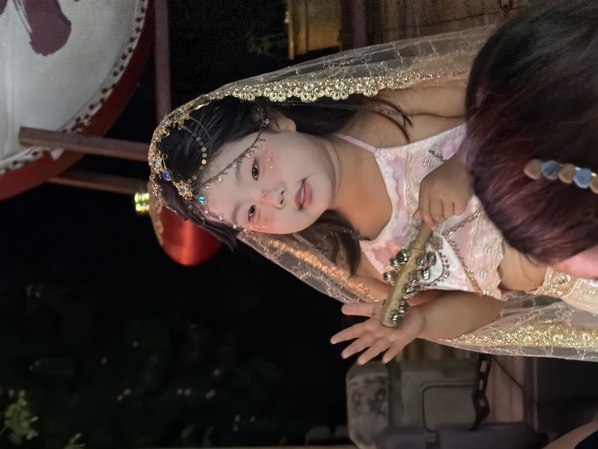 A young East Asian girl, appearing to be approximately 5 years old, is dressed in traditional ornate clothing and a veil. Her face is adorned with subtle makeup, including blush and sparkly accents near her eyes. She is holding a small, wooden object with metal jingles in her right hand, her fingers slightly spread. The girl is looking slightly to her left, with a gentle expression. She wears a bejeweled headpiece with hanging chains. Her attire consists of a light-colored halter top with ruffled trim and what appears to be a matching skirt or lower garment. A sheer, gold-trimmed veil drapes over her head and shoulders. The background is dark, suggesting it is nighttime, with some out-of-focus elements including what looks like a large, red, drum-like object supported by wooden beams in the upper right, and a red lantern visible to the left. The overall setting appears to be an outdoor event or performance in Yuzhong District, China.