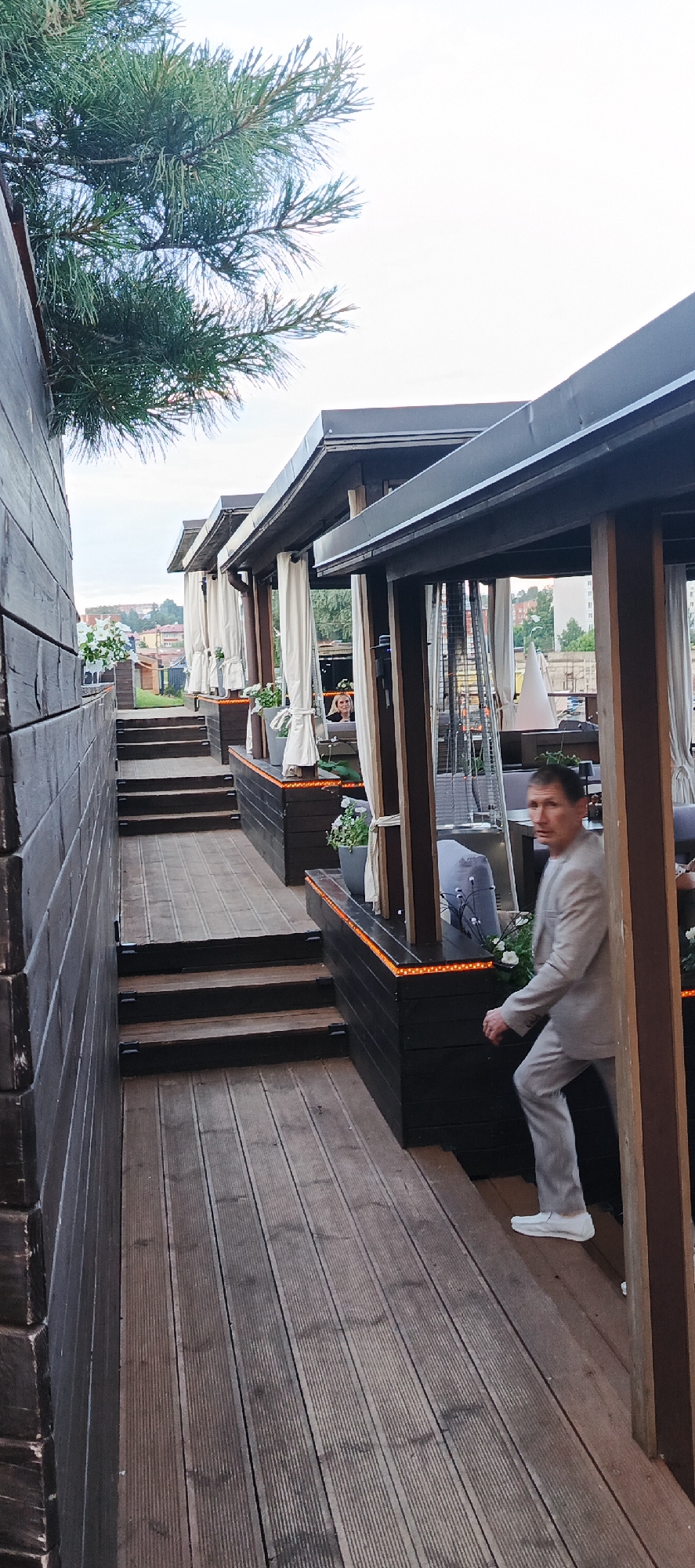 The image depicts an outdoor seating area of a restaurant or cafe, likely in Votkinsk, Russia, given the context provided. The area features multiple elevated wooden decks with stairs leading up to them. Each deck is covered by a canopy with white curtains. Decorative orange lighting runs along the base of the wooden structures. Potted plants and outdoor heaters are visible throughout the seating area.
In the foreground, a man in a light-colored suit and white shoes is ascending a wooden staircase, looking towards the viewer. Further back, a woman is visible seated at a table within one of the covered seating areas, interacting with a laptop. The background shows a slightly overgrown landscape with trees and some buildings in the distance, suggesting an urban or semi-urban setting. A pine tree branch hangs over the top left of the frame. The overall atmosphere appears to be that of a relaxed, upscale outdoor dining establishment.