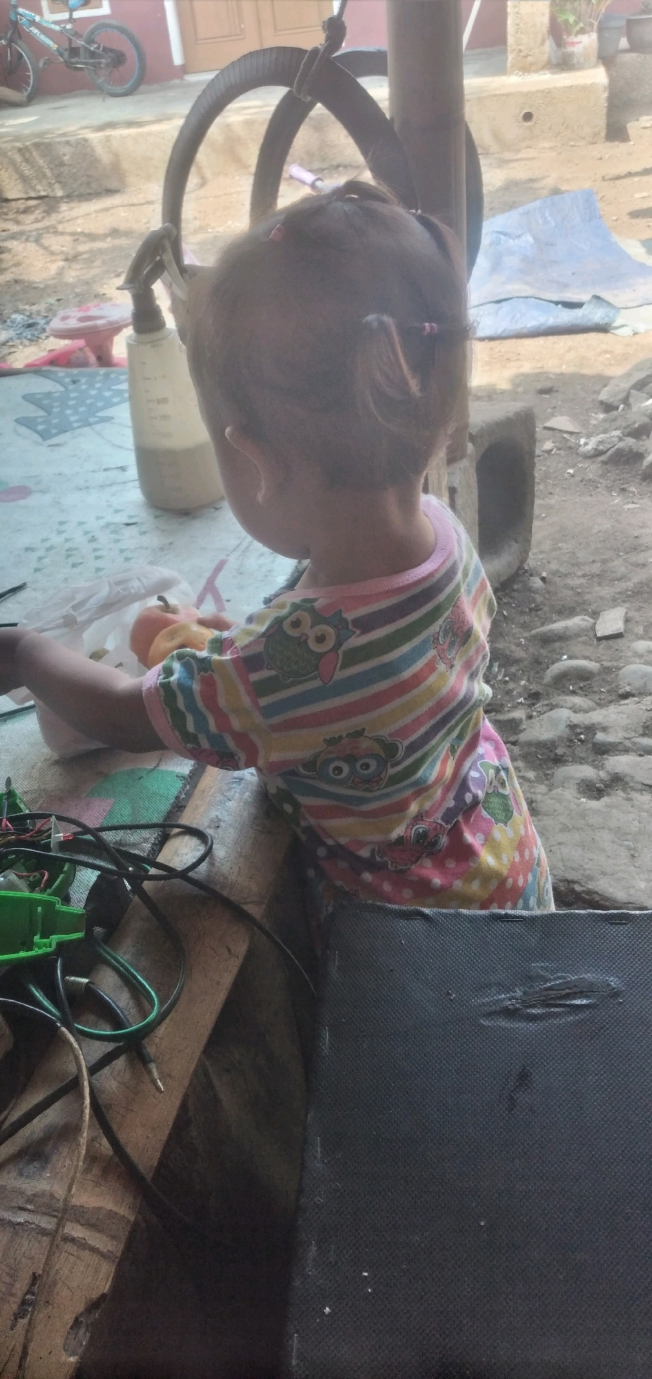 The image shows a close-up, rear-view of a young child with dark hair pulled into two pigtails, wearing a colorful striped shirt adorned with owl motifs. The child is seated at a wooden table, with their right arm extended and holding what appears to be an orange fruit. In front of the child, on the table, is a white plastic bag and various wires and electronic components, including a green plastic device. The background suggests an outdoor setting in Rahong, Indonesia, with rough, dirt ground, some scattered stones, and a rough concrete structure. A large, curved metal object hangs from above, partially obscuring the child's head, and a plastic bottle with markings is visible to the child's left. A bicycle is visible in the upper left portion of the frame. The overall scene is one of an informal, possibly domestic, outdoor environment.