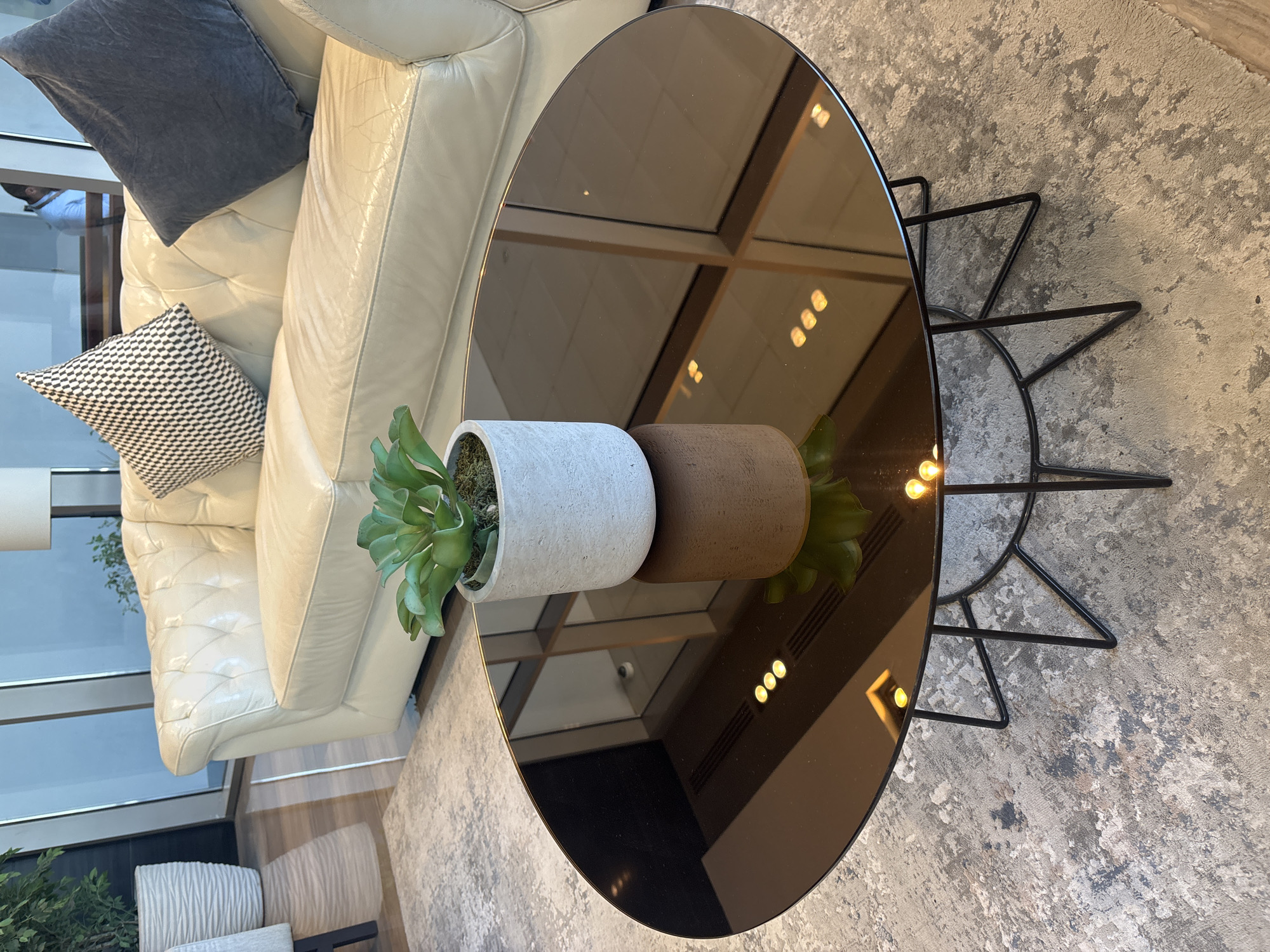The image shows a close-up view of a round, dark-brown, reflective coffee table with a modern black metal base. A small potted succulent plant sits on the table. In the background, a plush cream-colored tufted sofa is visible with two decorative pillows, one black and white patterned and the other dark gray. The environment appears to be a well-lit indoor space, possibly a lounge or waiting area. Reflections on the coffee table suggest a large window or glass partition, and possibly overhead lighting. Based on the quality of the furnishings and the overall aesthetic, this scene could be located in a high-end hotel lobby, a corporate office reception area, or a luxury residential building in Dubai, United Arab Emirates. There are no people clearly visible in the immediate foreground or middle ground of the image.
