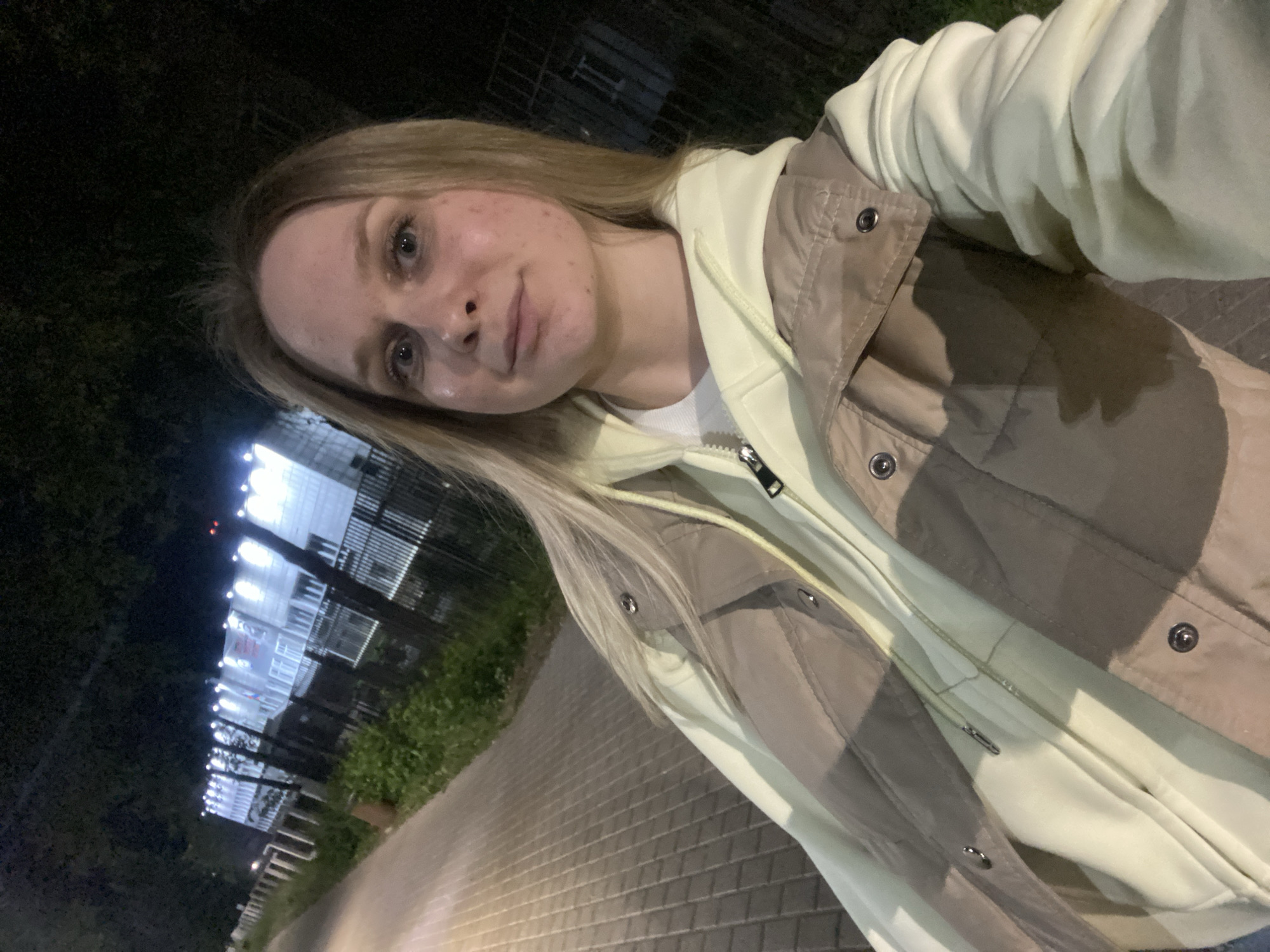 A young woman with blonde hair is taking a selfie outdoors at night. She is wearing a light yellow hoodie and a tan vest. Behind her, a sidewalk is illuminated by streetlights, and a building with numerous bright lights is visible. The building appears to be a school or administrative building with a fence in front of it. The surrounding area is dark with trees and greenery. The location is the city of Chaikovsky, Russia.