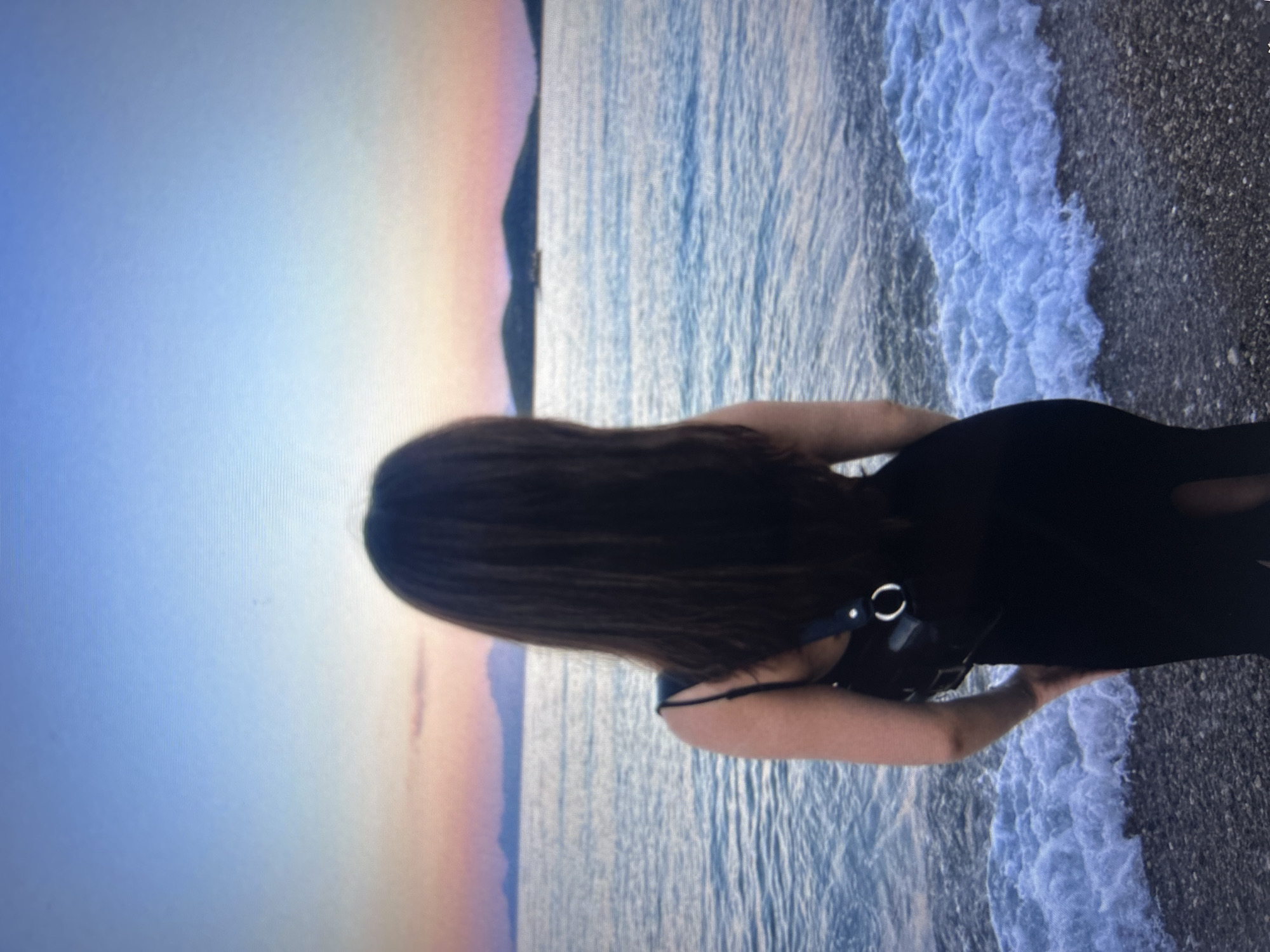 A person stands with their back to the viewer on a beach. The person has long, dark hair and is wearing a dark-colored outfit with thin straps. A strap with a circular object is visible on their back. Waves are washing onto a dark-colored, pebbly beach. In the background, the sky has hues of blue and pink, suggesting either sunrise or sunset. A landmass is also faintly visible in the background. The location cannot be determined in the city of Sofia, Bulgaria, as there is no beach in Sofia. The scene is a beach, possibly at sunset or sunrise.