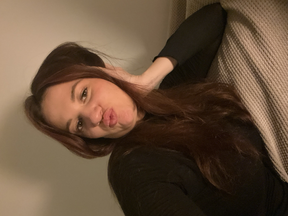 A woman with long, auburn hair poses with puckered lips, resting her head on her hand and arm. She wears a black, long-sleeved top. She is partially leaning against a textured, beige surface like a sofa or wall. The background is a plain, light-colored wall, and the lighting appears soft and diffused. The overall composition has a relaxed and casual feel.