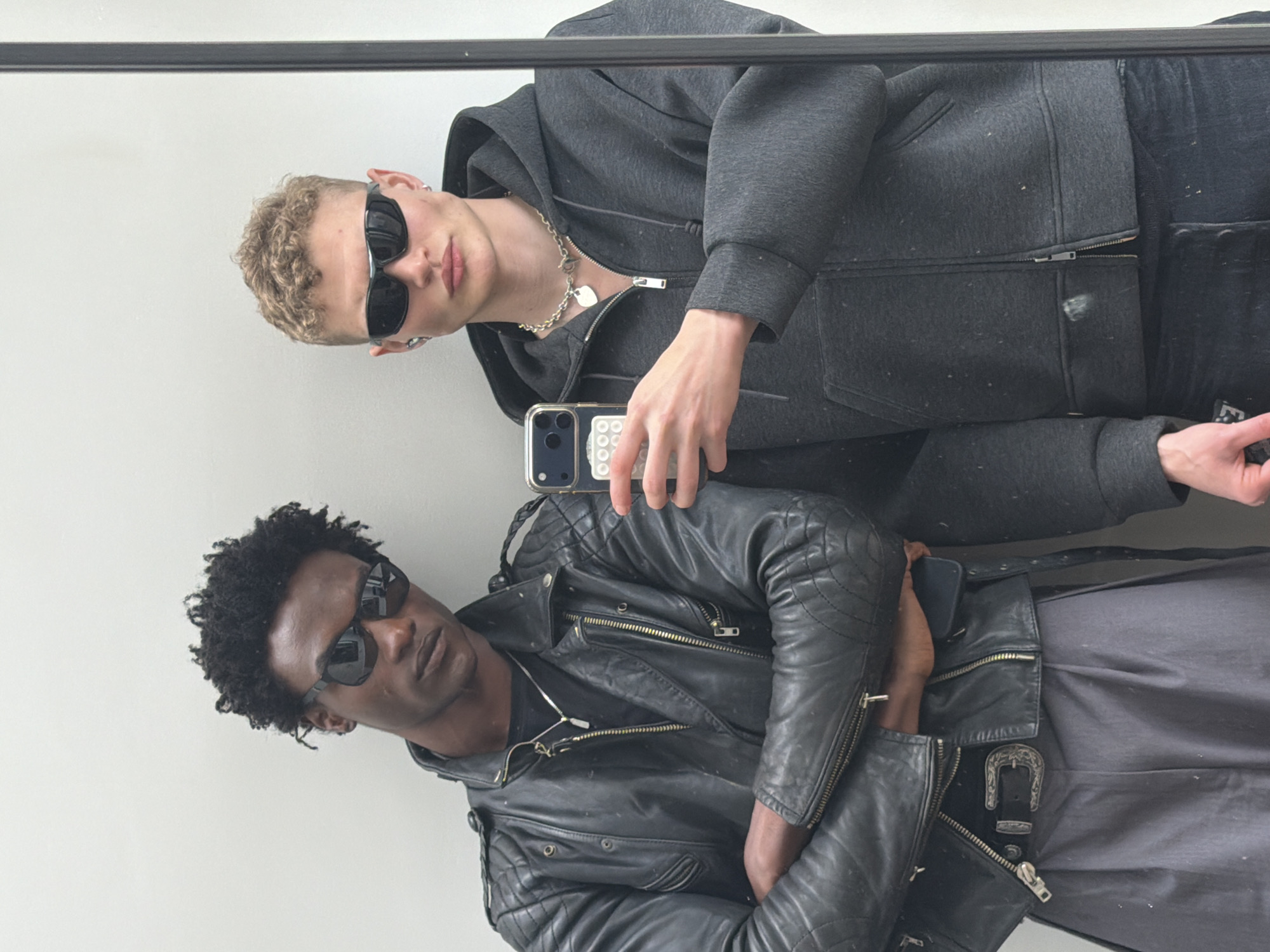 Two men are visible in the image, likely a mirrored reflection. The man in the foreground, facing the camera, has dark skin, short curly dark hair, and is wearing sunglasses, a silver chain necklace, and a black leather motorcycle jacket with prominent zippers and quilted detailing. A belt with a decorative buckle is visible at his waist. The man in the background has light skin, blond curly hair, and is also wearing sunglasses and a silver chain necklace. He is wearing a gray hooded sweatshirt and holding a smartphone. The environment appears to be indoors against a plain white wall. A dark horizontal line is visible at the top of the image, possibly a frame or edge of the mirror. The location is not discernible from the image.