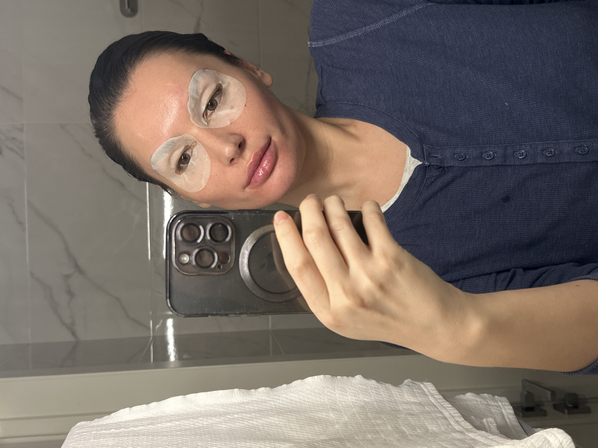 A person, appearing to be an adult female, is captured in a mirror selfie taken indoors, likely in a bathroom in Moscow, Russia. Her dark hair is pulled back, revealing her forehead. She has white, round eye masks positioned under each eye, suggesting a skincare routine. Her lips are glossy, and she wears a neutral to slightly smiling expression.
She is dressed in a dark blue, long-sleeved Henley shirt with several buttons down the placket, layered over a lighter undershirt. Her right hand holds a smartphone, visible in the mirror's reflection, indicating she is taking the photograph. The phone's camera array is prominent.
The background shows a light-colored tiled wall with a subtle marbled pattern, typical of a modern bathroom. Below the mirror, a white counter or ledge is visible, with a folded white paper towel or cloth resting on it. The image is oriented sideways, presenting the scene at an angle. No specific time of day can be determined from the indoor lighting, and there is no visible text in the image.