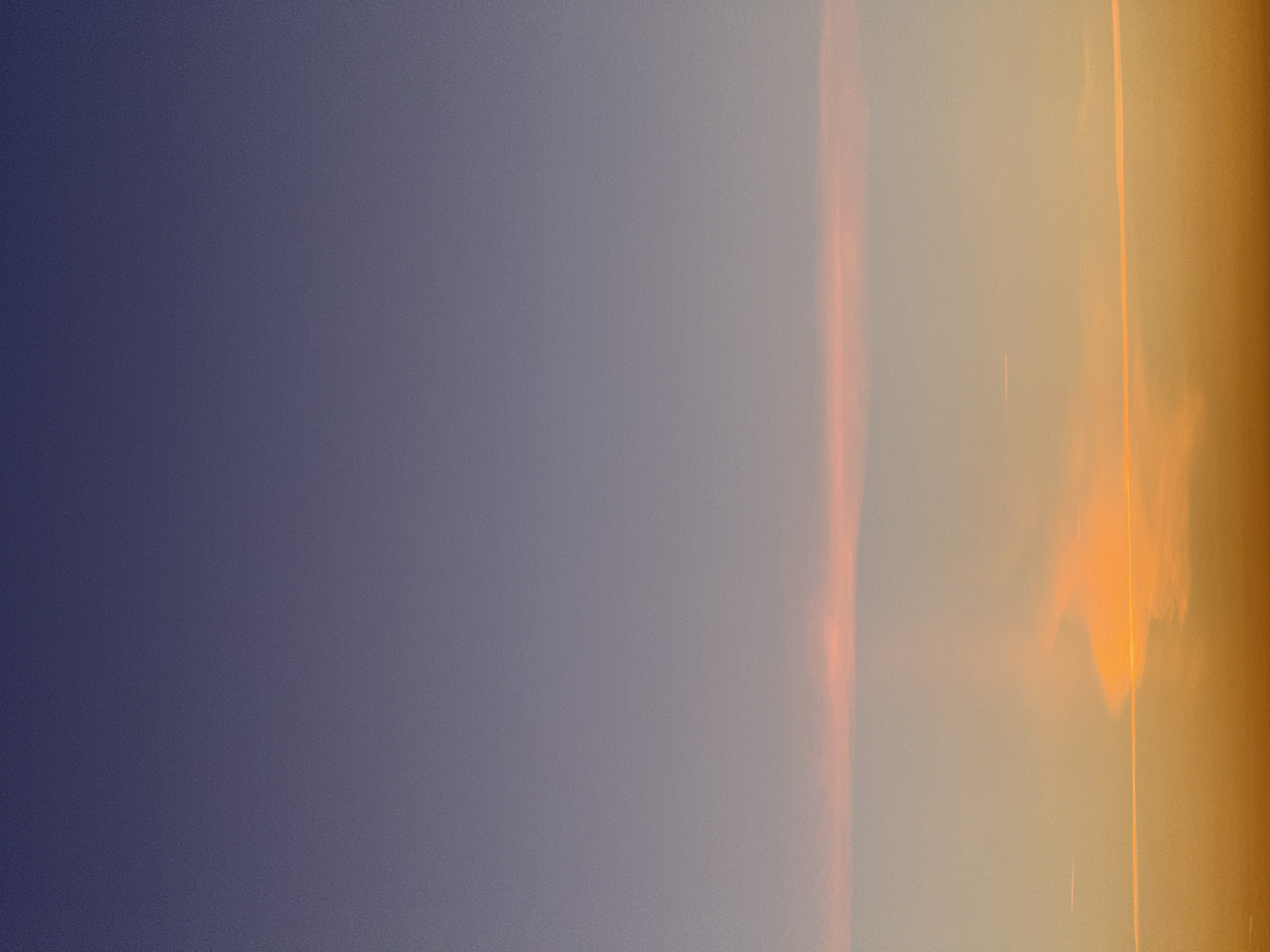 This is a vertical, full-frame photograph of a sunset sky over La Teste-de-Buch, France. The sky is a gradient of colors, transitioning from a deep indigo at the top to a warm orange at the bottom. There are faint, thin clouds present, with one prominent band of pinkish-orange clouds stretching horizontally across the middle of the frame. Several faint contrails from aircraft are visible as thin white lines against the colored sky. The overall scene is serene and devoid of any human presence or discernible man-made structures.