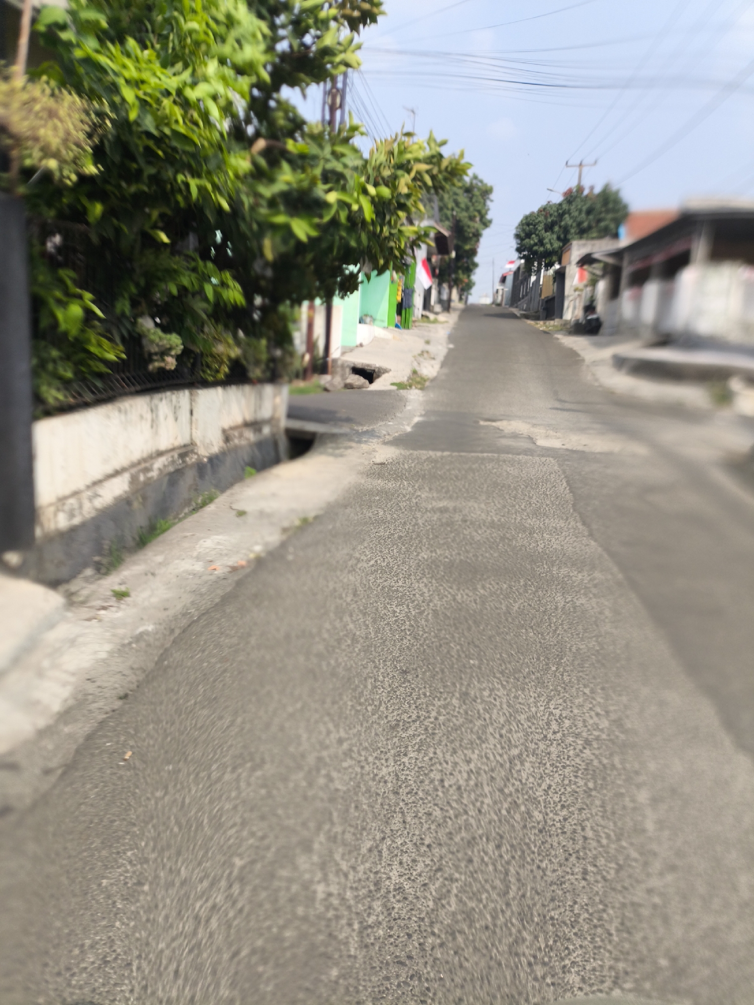 The image captures a narrow, paved road in a residential area. The road ascends slightly into the distance. On the left side of the road, there is a concrete wall with a metal fence on top, behind which are lush green trees and foliage. A drainage ditch runs alongside the road on the left. On the right side of the road, there are several low-rise buildings with varied architectural styles and colors, suggesting a dense urban or semi-urban environment. Power lines are visible crisscrossing the sky above. The lighting indicates it is daytime with clear weather. The overall impression is of a typical street scene in a neighborhood.
