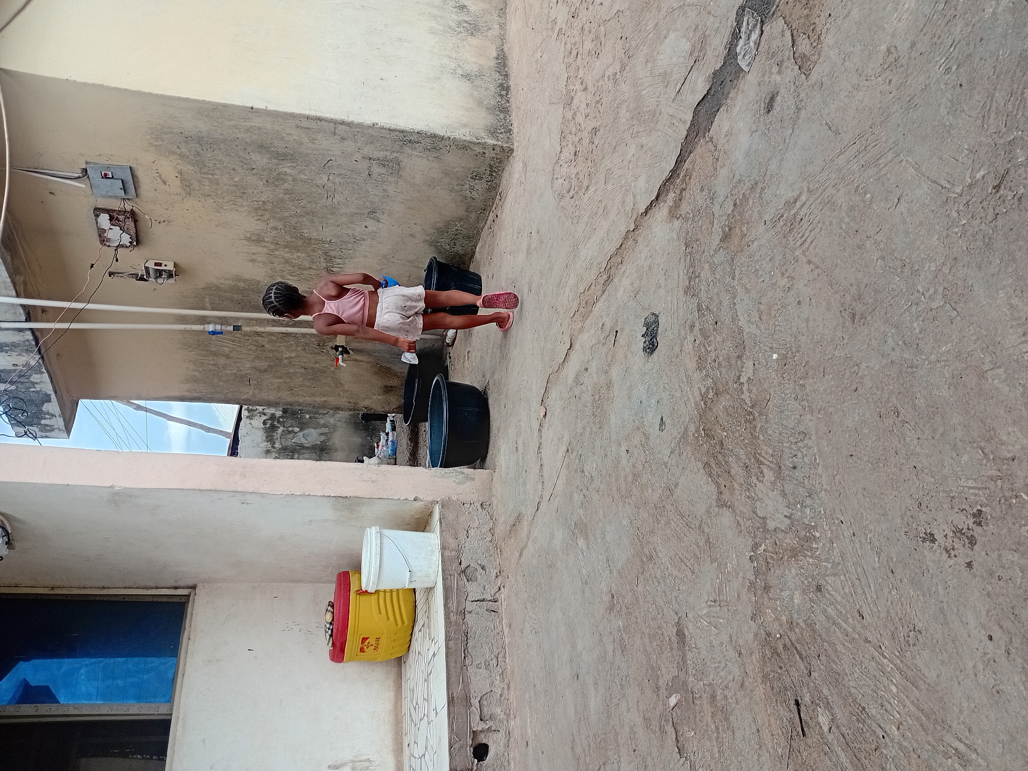 The image depicts a young girl standing in an outdoor space. The girl is wearing a pink top, white shorts, and red sandals. She appears to be washing something, with a blue cloth in her hand, near several large black buckets. The surrounding environment is a concrete courtyard or alleyway, bordered by walls. One wall has electrical fixtures and exposed wiring. Two buckets, one yellow and one white, are stacked next to a wall. The scene is situated in the city of Unknown, Unknown.