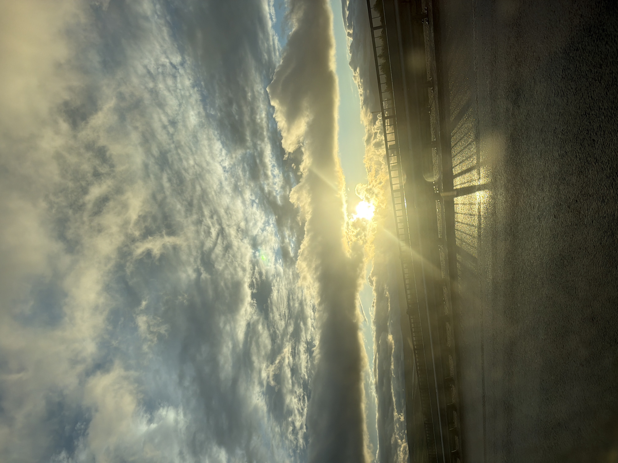 This media file is an outdoor photograph, presented in a vertical orientation, but depicting a landscape scene likely viewed horizontally. It captures a dramatic moment, likely at sunrise or sunset, over what appears to be a wet road or bridge. The location context provided is Blatino, Bulgaria.
The left side of the frame (when viewing the scene horizontally) is dominated by a dynamic sky filled with a mix of clouds. These range from darker, textured grey formations to bright, luminous white and golden clouds, suggesting a complex weather pattern. Patches of lighter blue sky are visible in between the cloud masses.
On the right side of the frame, a brilliant sun is positioned low on the horizon, partially obscured by clouds. It emits intense sunbeams and a strong lens flare, bathing the surrounding clouds and atmosphere in warm, golden light.
A dark, linear structure, resembling a railing or fence, runs horizontally across the mid-section of the image, marking the horizon line. This structure separates the vibrant sky from the foreground surface.
Below this horizon line, the right half of the image features a dark, textured surface, likely asphalt, which appears wet and highly reflective. The wetness creates striking specular highlights and mirrors the bright sun and parts of the sky. The railing casts long, sharp, ladder-like shadows that stretch across this glistening surface, intensified by the low angle of the sun. The bright reflection of the sun itself is also prominent on the wet ground.
The overall impression is one of a serene yet powerful natural spectacle, characterized by the interplay of light, clouds, and reflections after a recent rain. No people or specific activities are visible, and there is no discernible text in the image.