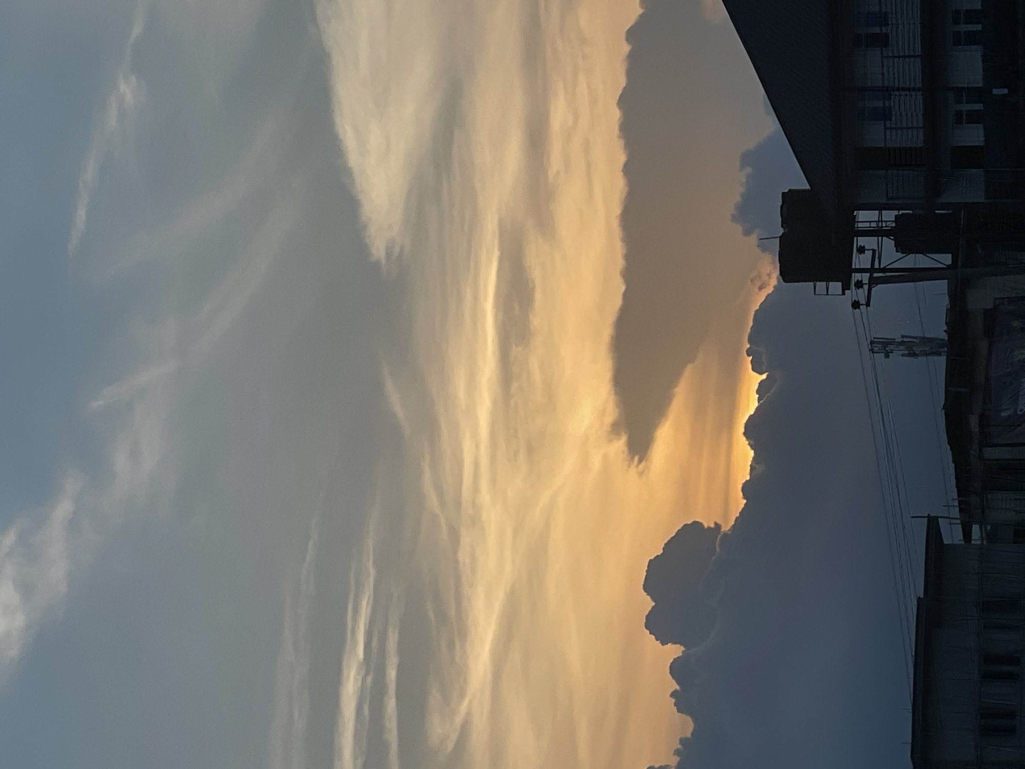 The image captures a dramatic sunset over the city of Onitsha, Nigeria. The sky is a canvas of vibrant oranges, yellows, and blues, with wispy clouds scattered across the upper portion and thick, dark clouds dominating the lower half. The setting sun peeks through a gap in the clouds, casting a warm glow. Silhouetted against this breathtaking sky are buildings, including a multi-story structure with a dark, pitched roof and balconies, and a water tank atop a metal frame. Power lines crisscross the scene, and a cell tower is visible in the distance. The overall impression is one of urban life set against a powerful natural spectacle.