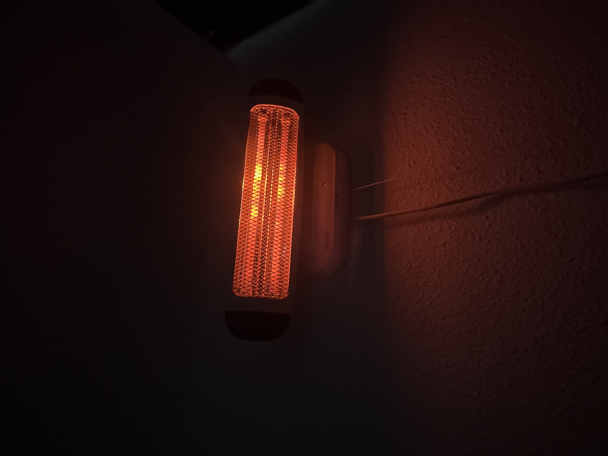An infrared heater is mounted on a dark, textured ceiling. The heater is rectangular with a wire mesh front that glows orange from the heat. A white power cord extends downwards from the heater. The surrounding environment is dark, with the light from the heater illuminating the textured surface below.