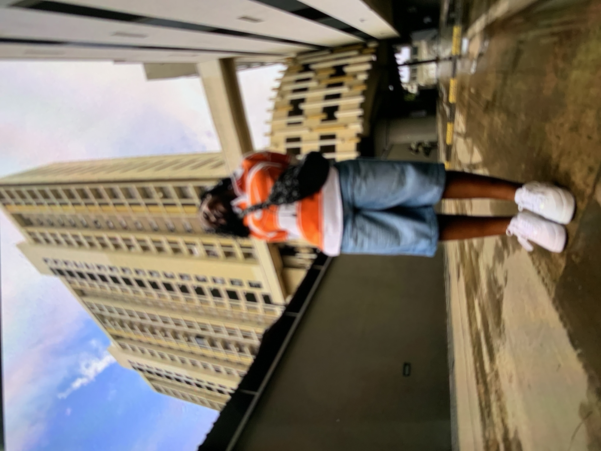 The image shows a person standing on a walkway with a tall building in the background. The building appears to be a multi-story structure with repetitive windows. The person is wearing shorts, a shirt, and white shoes, and has a bag across their chest. The walkway surface is textured and appears to be made of concrete. The photo is taken from a low angle.
The building in the background is likely the Tanzania Ports Authority Headquarters (TPA) in Dar es Salaam, Tanzania.
