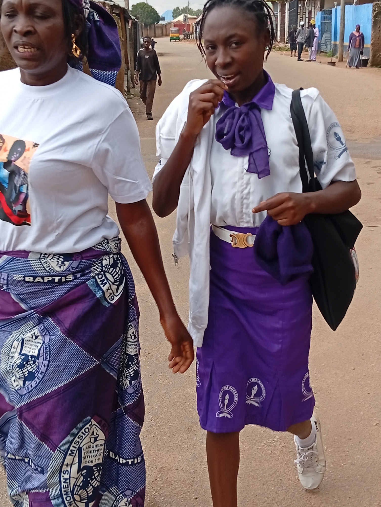 Two women, appearing to be of African descent, walk on a dirt road in Jos, Nigeria. The woman on the left, her face is partially visible, wears a white t-shirt with a picture of a person printed on it and a purple and white patterned skirt. The woman on the right, her face is fully visible, wears a white shirt with a purple ribbon tied at the neck and a solid purple skirt. She also carries a black bag over her shoulder. In the background, other people are visible walking and standing, with buildings and a yellow vehicle in the distance, suggesting a busy urban environment. The ground is unpaved, with some loose dirt and gravel. The overall scene depicts a street in a developing city, likely in West Africa.