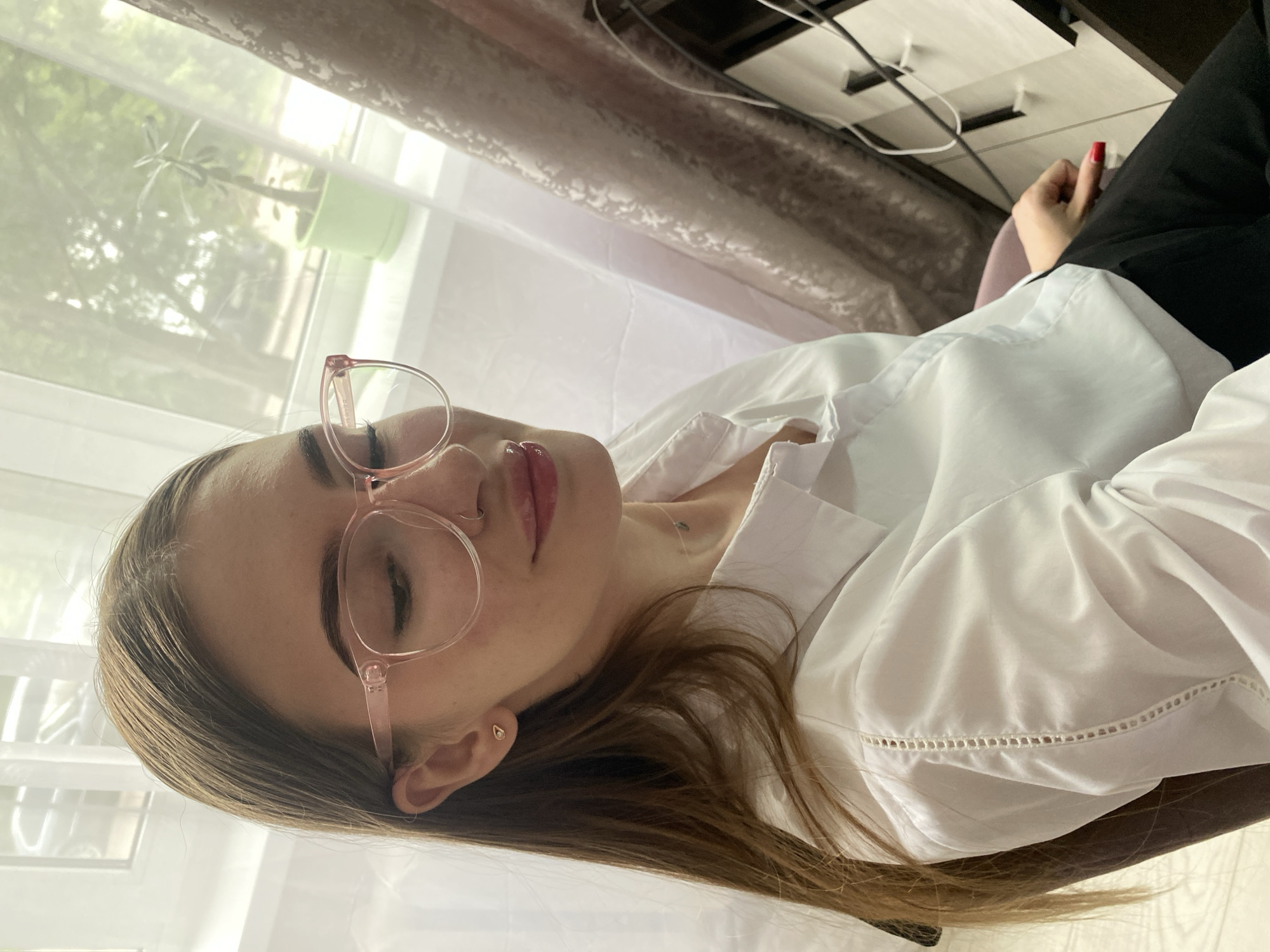 A young woman wearing pink-rimmed glasses, a white button-down shirt, and black pants is seated indoors. Her hair is long and brown, and she has on subtle makeup. She is looking down and to her right. A small, heart-shaped earring is visible in her left ear. Behind her, a window shows trees and what appears to be a plant in a green pot. To her right, part of a wooden cabinet with some wires is visible, along with a brown curtain. The woman's right hand rests on her lap, and her fingernails are painted red. The scene is likely a home or office environment.