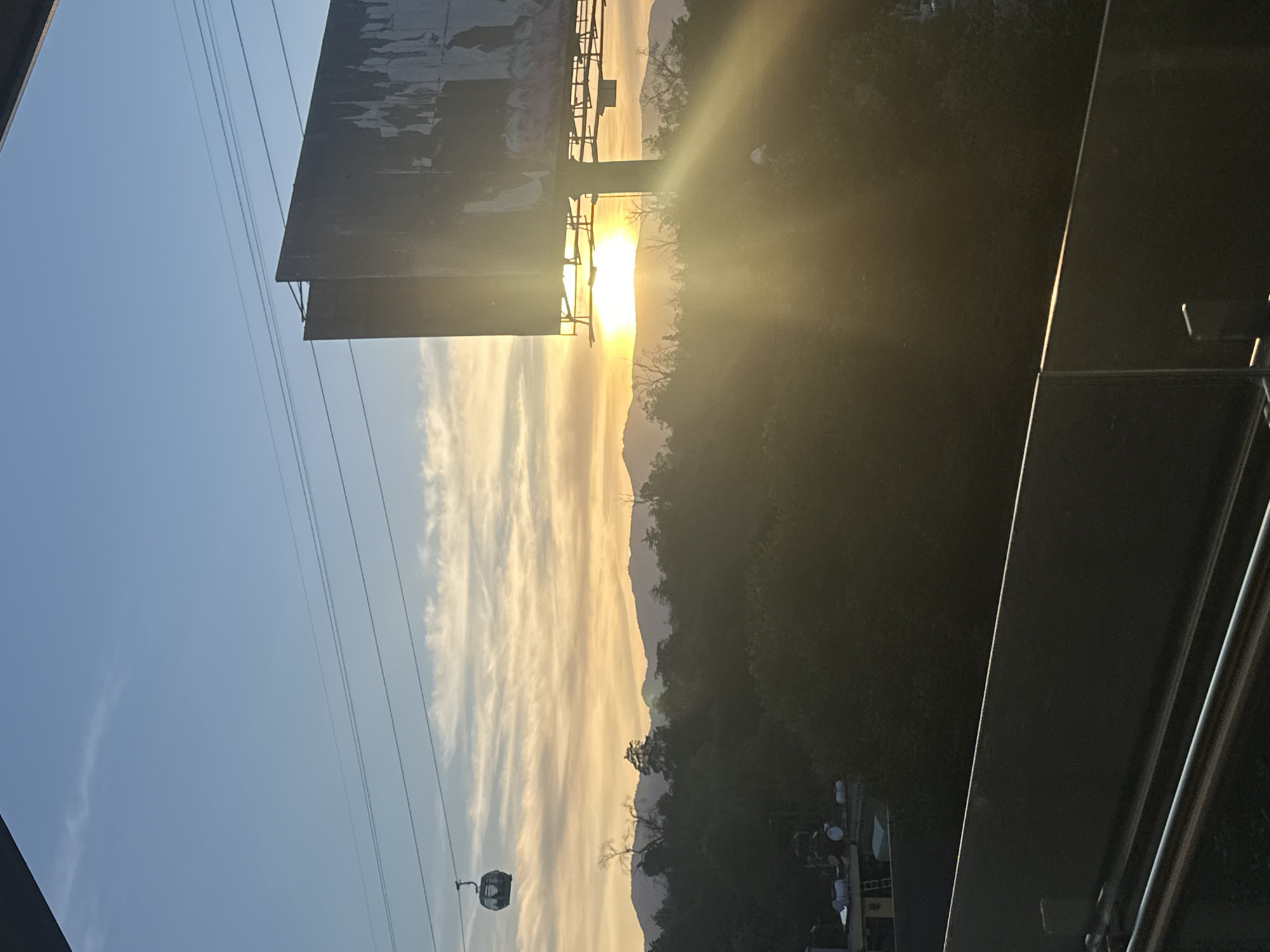 This is an outdoor scene taken in Mexico City. A sunset is obscured by a large billboard. There are mountains in the distance. A gondola and cable wires are on the left side of the frame. Vegetation is to the right of the frame. A railing and a partial building frame the right edge of the image.