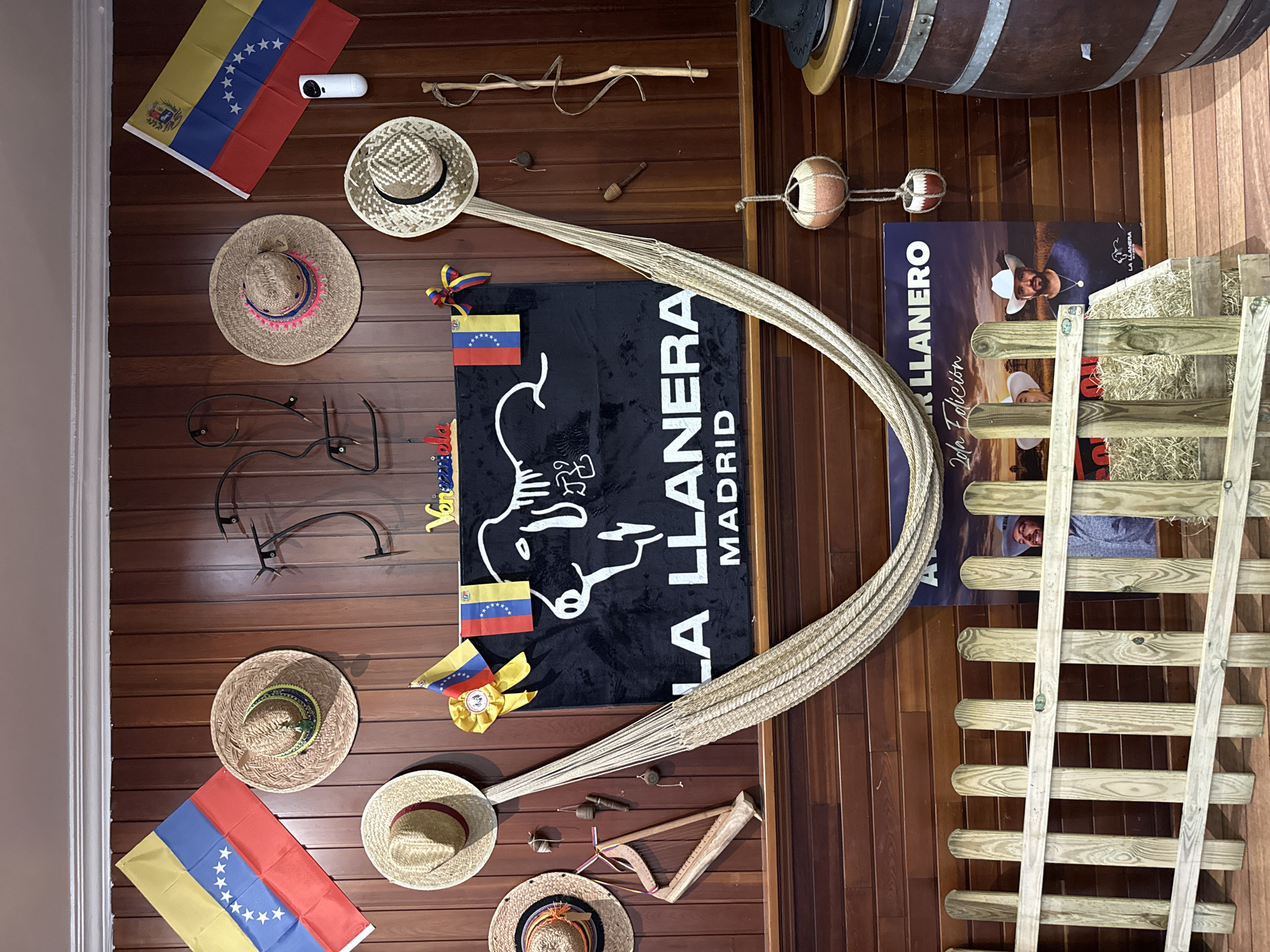 The image shows a wall decorated with Venezuelan flags, straw hats, traditional Venezuelan items like a small harp and spinning tops, and a large black banner with a white illustration of a cow and the text "LA LLANERA MADRID". A hammock is hung from the wall. In the lower part of the image, a wooden fence is in front of a poster featuring two men in cowboy hats. A barrel is visible on the right. The overall impression is a display of Venezuelan culture, possibly in a restaurant or cultural center. Torrejón de Ardoz, Spain is mentioned as the location of this establishment.