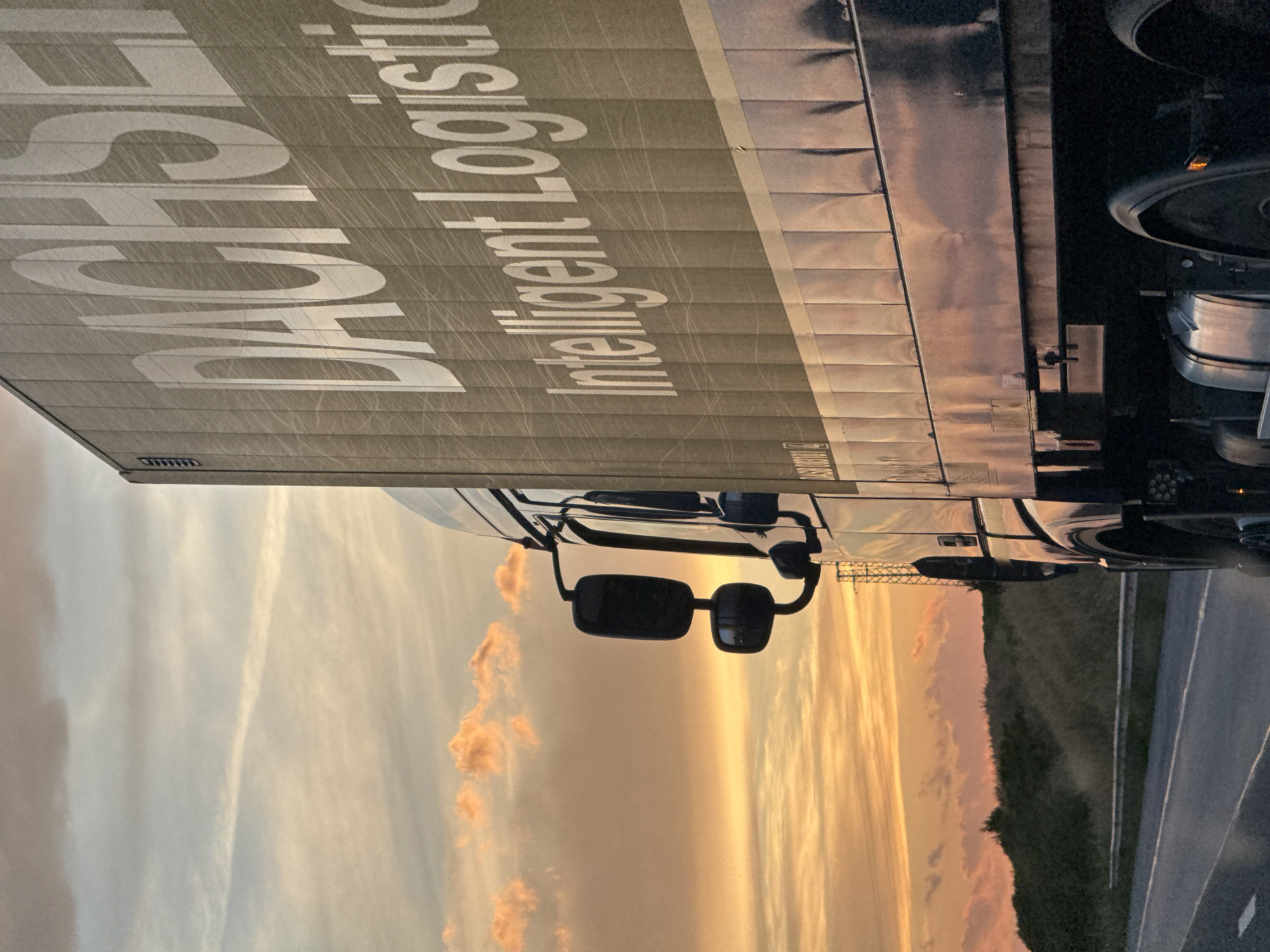 The image shows the side of a truck with "DACHSER Intelligent Logistics" written in large white letters. The truck is silver and appears to be a semi-trailer. The sun is setting, casting a warm glow over the sky and clouds, which are a mix of orange, pink, and white. The truck is parked on the side of a road, with a grassy embankment visible in the background. There are lane markings on the road, indicating it is a highway or a similar road. The overall mood is serene and peaceful, with the setting sun creating a beautiful backdrop for the industrial subject. The image is oriented vertically, as if the viewer is looking up at the truck.