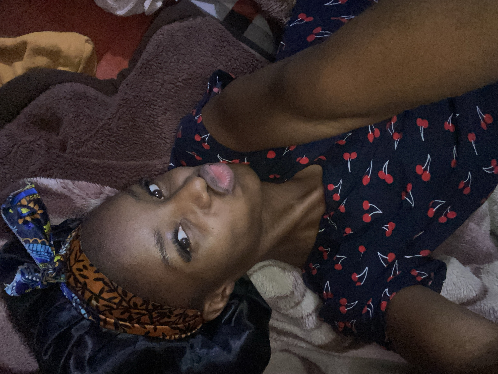 The image depicts an adult female positioned in a prone or semi-reclined orientation, taking a selfie indoors. Her complexion is dark. She has dark eyes and defined eyelashes, looking directly at the camera with her lips puckered into a 'kissing' pose. Her head is adorned with a patterned headscarf featuring orange-brown leaf-like motifs, overlaid with a multi-colored (blue, yellow, black) knotted fabric. A dark satin bonnet is visible underneath the headscarf on her left side (viewer's right). She is wearing a dark blue garment patterned with red cherries and white stems. Her left arm (viewer's right) extends towards the camera, suggesting she is holding the device. She is situated on soft bedding or textiles; a light-colored, possibly textured fabric is beneath her left side, and a dark brown, plush fabric is to her right. The background implies an interior space with muted lighting. This scene is situated in Jos, Nigeria.