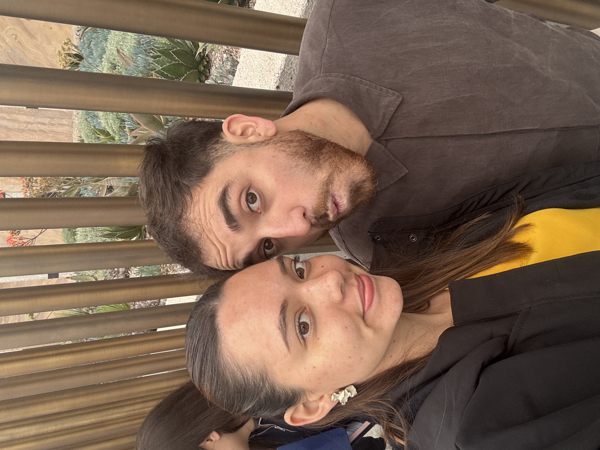 Two individuals, a man and a woman, are captured in a close-up, likely selfie-style portrait. The man, positioned on the left, has dark hair, a beard, and brown eyes, wearing a dark gray collared shirt. He looks towards the viewer with a neutral, slightly smirking expression. The woman, on the right, has long dark brown hair, brown eyes, and fair skin with freckles. She is wearing a black top over a yellow layer and a distinctive white, wavy earring on her right ear, smiling gently at the camera. A hint of another person's head is visible in the bottom left corner, out of focus.
The subjects are outdoors, bathed in bright, natural daylight, suggesting a clear day. Behind them, a modern metal slatted railing or barrier is visible. Through the gaps in the railing, a lush landscape of various succulent plants, including agaves and possibly flowering aloes, can be seen, indicating a botanical garden or a carefully curated outdoor space. The setting, combined with the provided context of Mexico City, Mexico, suggests a location like the Jardín Botánico del Bosque de Chapultepec or a similar arid plant collection. Both individuals appear relaxed and content, posing together for the photograph. No text is visible in the image.