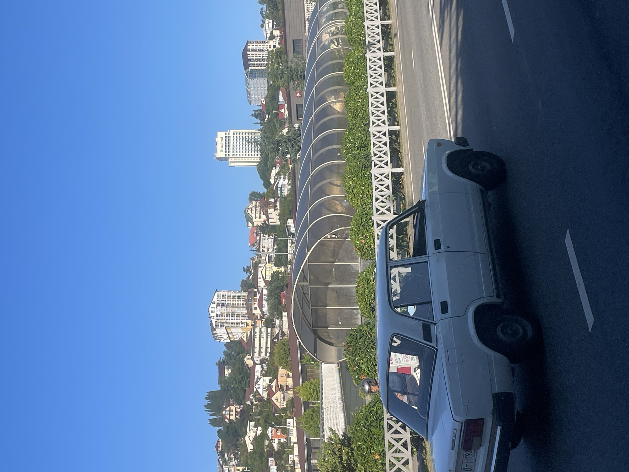 The image depicts a sunlit street scene in Sochi, Russia. In the foreground, a white Lada sedan is driving on the right side of a road. The car's license plate is partially visible, showing "60TT 193". A person wearing a helmet is seated in the driver's seat. Behind the car, a white decorative fence lines the road, separating it from a green, manicured area and a long, arched, transparent structure that appears to be a covered walkway or greenhouse. Further in the background, a hillside is covered with numerous buildings, ranging from smaller houses to multi-story apartment complexes and a tall white hotel with the "AZIMUT" logo. The sky is a clear, bright blue. The overall impression is of a coastal city on a clear day.