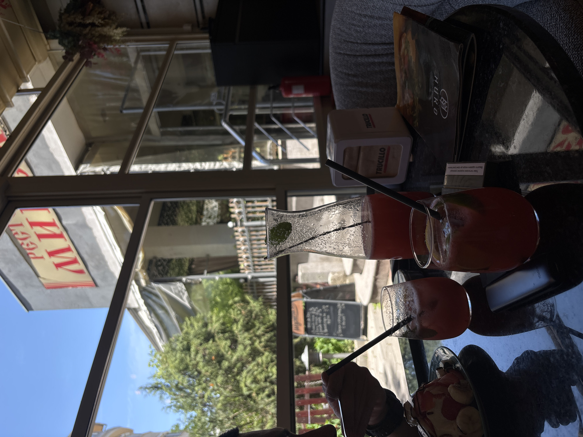 The image shows a table at an outdoor cafe in Plovdiv, Bulgaria, with a view of a building with "РЕСТ МИ" signage. On the table, there are two glasses of red-colored drinks with straws and mint leaves, a glass pitcher filled with the same drink, a napkin holder with "TRUCILLO" branding, a cigarette pack, a menu, and a plate with a dessert topped with fruit. A person's hand is visible holding a straw. The background shows trees and other cafe elements through a window.