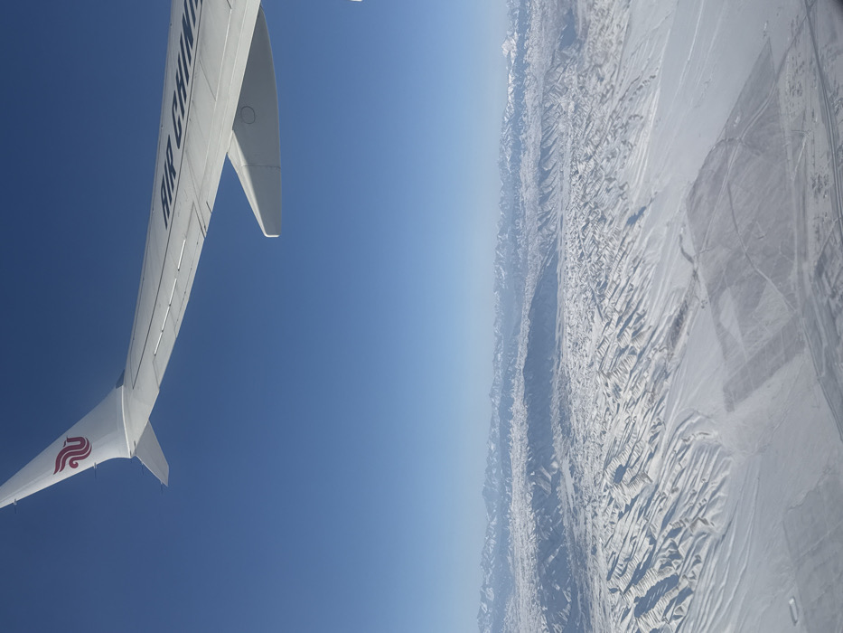 An aerial view from an aircraft window captures a vast, snow-covered landscape under a clear, deep blue sky, identified as being over the region near Sangong, China. The white wing and a portion of the tail fin of an Air China aircraft, bearing its red phoenix logo and partial "AIR CHINA" text, are visible in the upper left quadrant. The terrain below consists of undulating, snow-blanketed hills and valleys in the mid-ground, displaying sharp contrasts between sunlit surfaces and shadows. In the distant background, a long range of snow-capped mountains stretches across the horizon. Flatter, snow-covered areas with faint linear patterns, possibly indicating fields or infrastructure, are discernible in the lower right portion of the frame. No individuals are visible. The scene depicts an aerial flight over a wintry, mountainous environment.