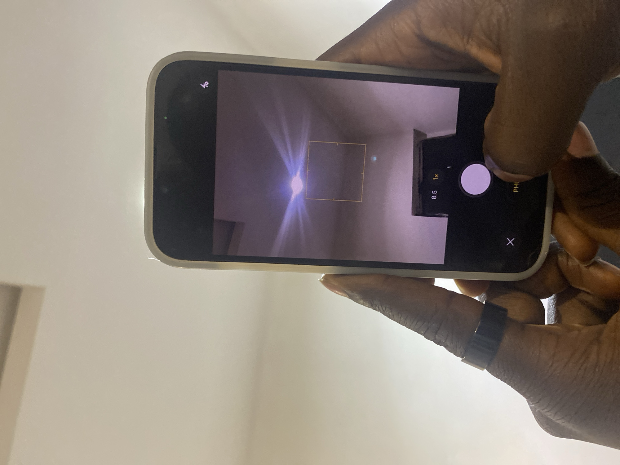 The image shows a close-up of two hands holding a smartphone. The smartphone is positioned to capture a ceiling light fixture with a square focus box superimposed on it. The phone screen displays camera interface elements, including zoom options (0.5x and 1x) and a large circular capture button. One hand wears a black ring on its index finger. The background is a plain, light-colored ceiling. There is no visible indication of the city of Jos, Nigeria in the image itself.