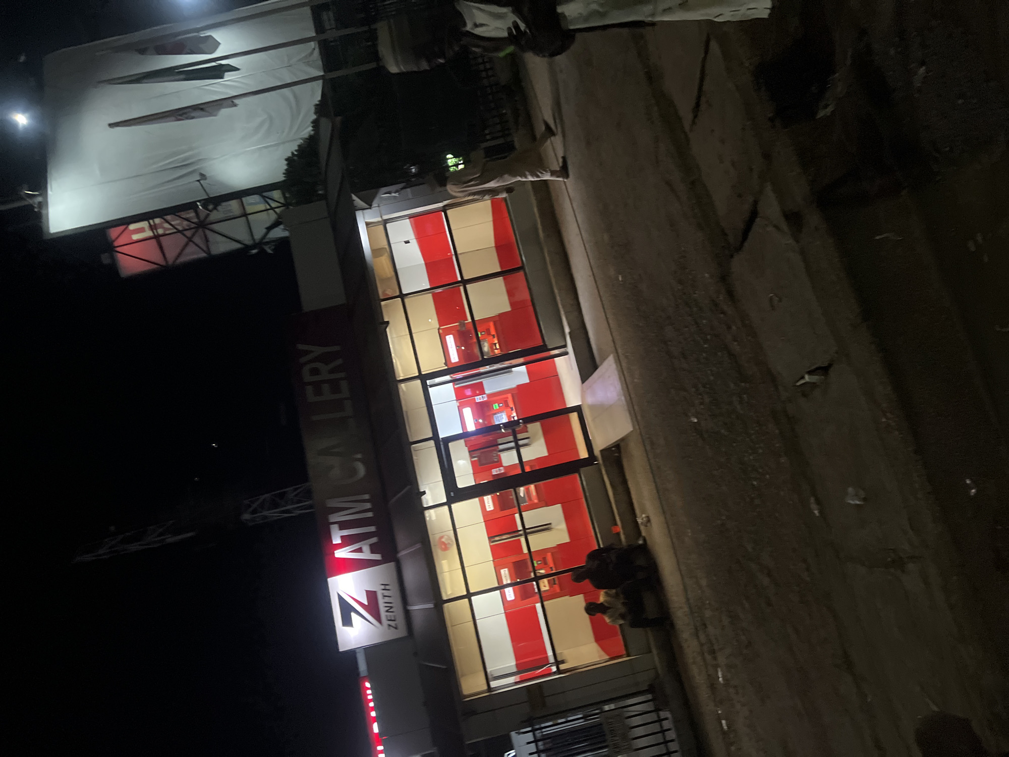 This nighttime photograph depicts a Zenith Bank ATM Gallery located in the city of Jos, Nigeria. The gallery's exterior features a prominent "Zenith ATM Gallery" sign with the bank's logo. The interior is brightly lit, revealing a red and white color scheme and multiple ATM machines through a large glass facade.
Two individuals are seated on a low step or curb directly in front of the ATM gallery, to the left of the main entrance. A third individual, dressed in light-colored traditional attire, walks away from the gallery towards the right side of the frame. On the far right, partially obscured, a fourth person in similar attire stands, holding a mobile device and facing towards the left.
The foreground consists of a dark, unpaved or roughly paved ground surface with scattered debris. To the left, a dark metal fence or gate is visible. In the background, on the left, a tall, dark communication tower or mast rises against the night sky. On the right, a large billboard structure with a white canvas is prominently illuminated, and three flags are visible on poles in front of it. Bright light sources are visible in the upper right quadrant of the image.