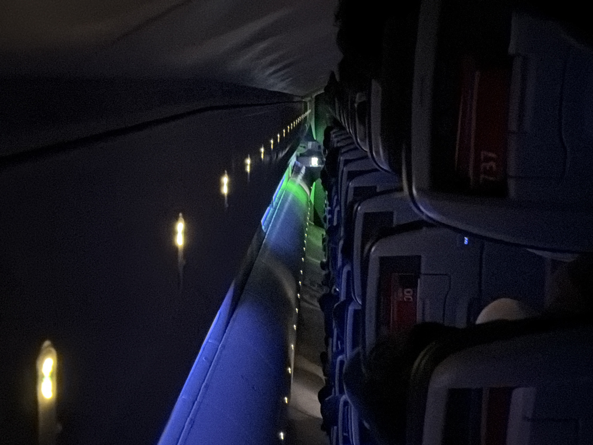 The scene is the interior of a Boeing 737 airplane. The cabin lights are dimmed, and aisle lights illuminate the walkway with a blue-green hue. Seats line the right side of the frame. Seat numbers, such as '30' and '737,' are visible on the seat backs. Passengers are visible sitting in the seats, their heads and shoulders silhouetted against the dark background. The location cannot be determined to be within the city of Los Angeles, United States of America.