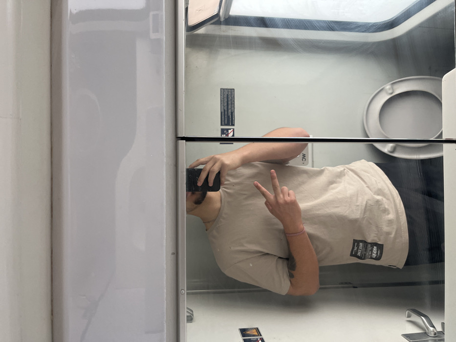 A male individual is reflected in a mirror, taking a selfie inside a confined space consistent with a train lavatory, located in Stambolovo, Bulgaria. The subject, seen from the chest up, has a short beard and is wearing a plain, light beige short-sleeved t-shirt. A black smartphone with a cracked screen protector is held in his right hand, used for taking the photograph. His left hand is raised, displaying a "peace sign" with the index and middle fingers. A pink bracelet is visible on his left wrist, and a dark tattoo is partially visible on his left forearm. In the background reflection, a white toilet with an open lid is positioned on the right side. To the left, a light-colored wall features a metallic door handle and two small warning signs. On the right, beyond the toilet, a window with streaked glass shows an indistinct exterior. The overall environment features light-colored walls and overhead panels.