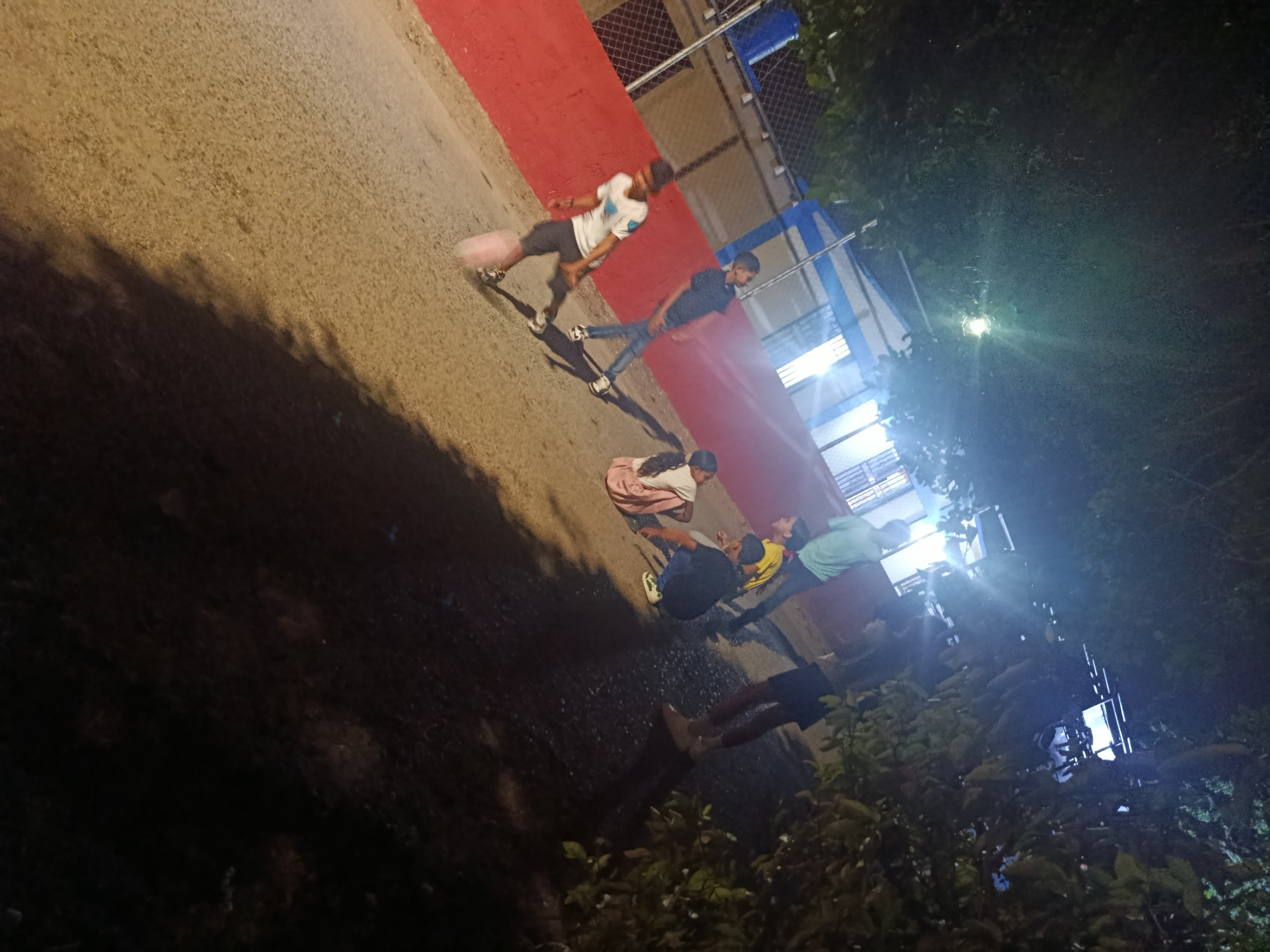 At night in Juan Griego, Venezuela, a group of people stand on a concrete walkway adjacent to a red wall and metal fencing. A streetlight illuminates the scene. The environment includes trees and foliage, contributing to the darkness of the background. The subjects appear to be interacting with one another.