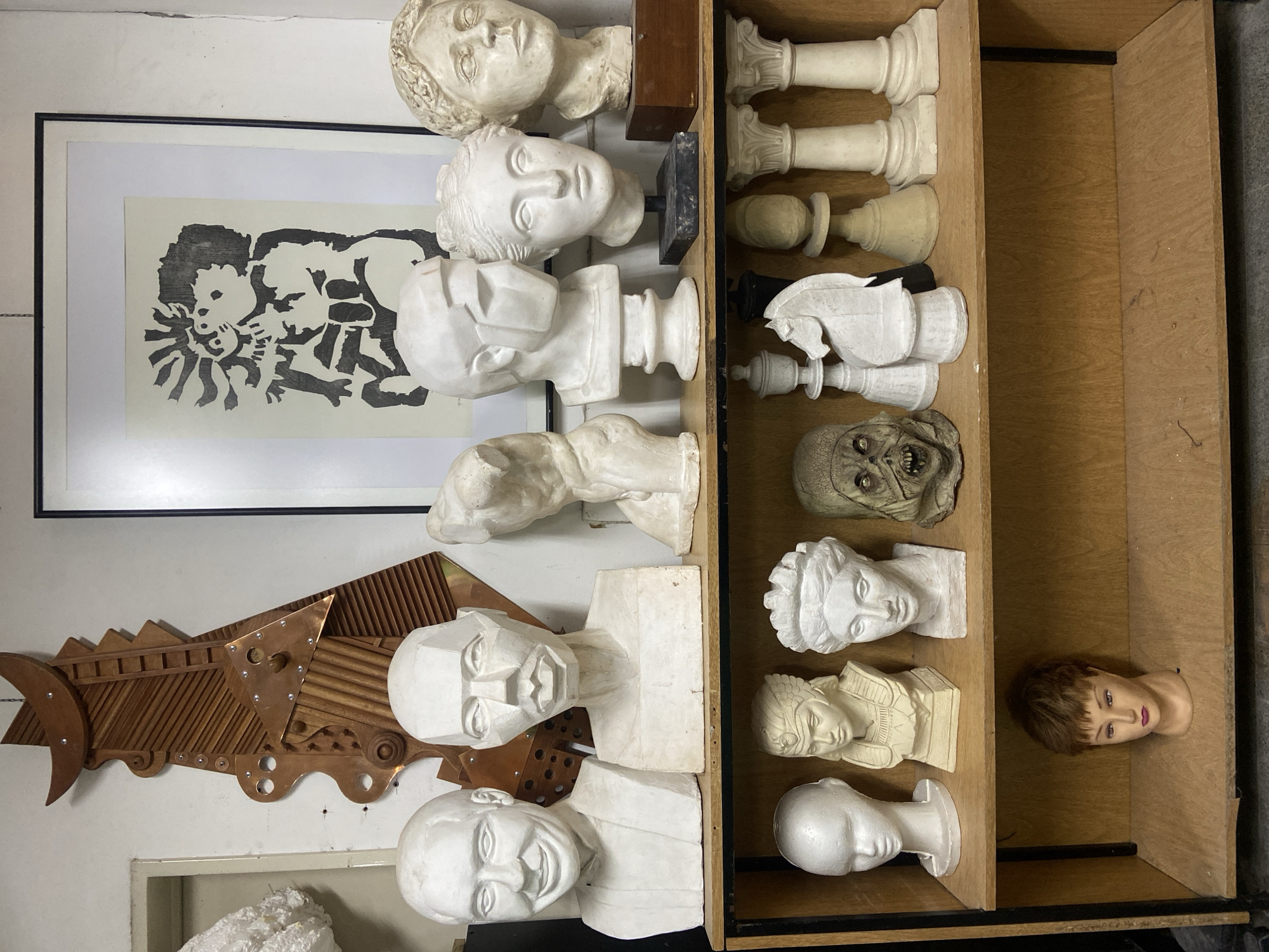 This media file captures a vertical view of an indoor space, likely an art studio, workshop, or storage area, located in Buenos Aires, Argentina. The scene is filled with a diverse collection of sculptures, busts, and art pieces displayed on shelves and mounted on white walls.
On the left, a large, intricate wall sculpture crafted from wood and metallic elements features geometric shapes, ribbed textures, and shiny fastenings. Below it, several white plaster or ceramic busts are arranged, including a cubist-inspired head, a smiling male bust, and other human head forms with distinct anatomical details.
A prominent vertical wooden shelving unit on the right side houses a variety of items. The upper shelves are occupied by large, white chess pieces, such as pawns, rooks, and knights, all uniformly crafted from a material resembling plaster. Below these, a greenish-grey skull or mask with a grotesque, open-mouthed expression and visible teeth is positioned. Further down the shelves, more white plaster busts are displayed, depicting classical female heads, a bust in an ancient Egyptian style, and a sleek, minimalist head sculpture. On the bottom shelf, a female mannequin head with short brown hair and makeup lies on its side.
In the upper left portion of the image, a framed black and white print is visible, featuring stylized, abstract figures in a dynamic composition. The scene is static, showing a collection of artistic creations and anatomical models rather than an active event. The environment suggests a creative workspace or a curated collection of sculptural forms. No discernible text is visible within the image.