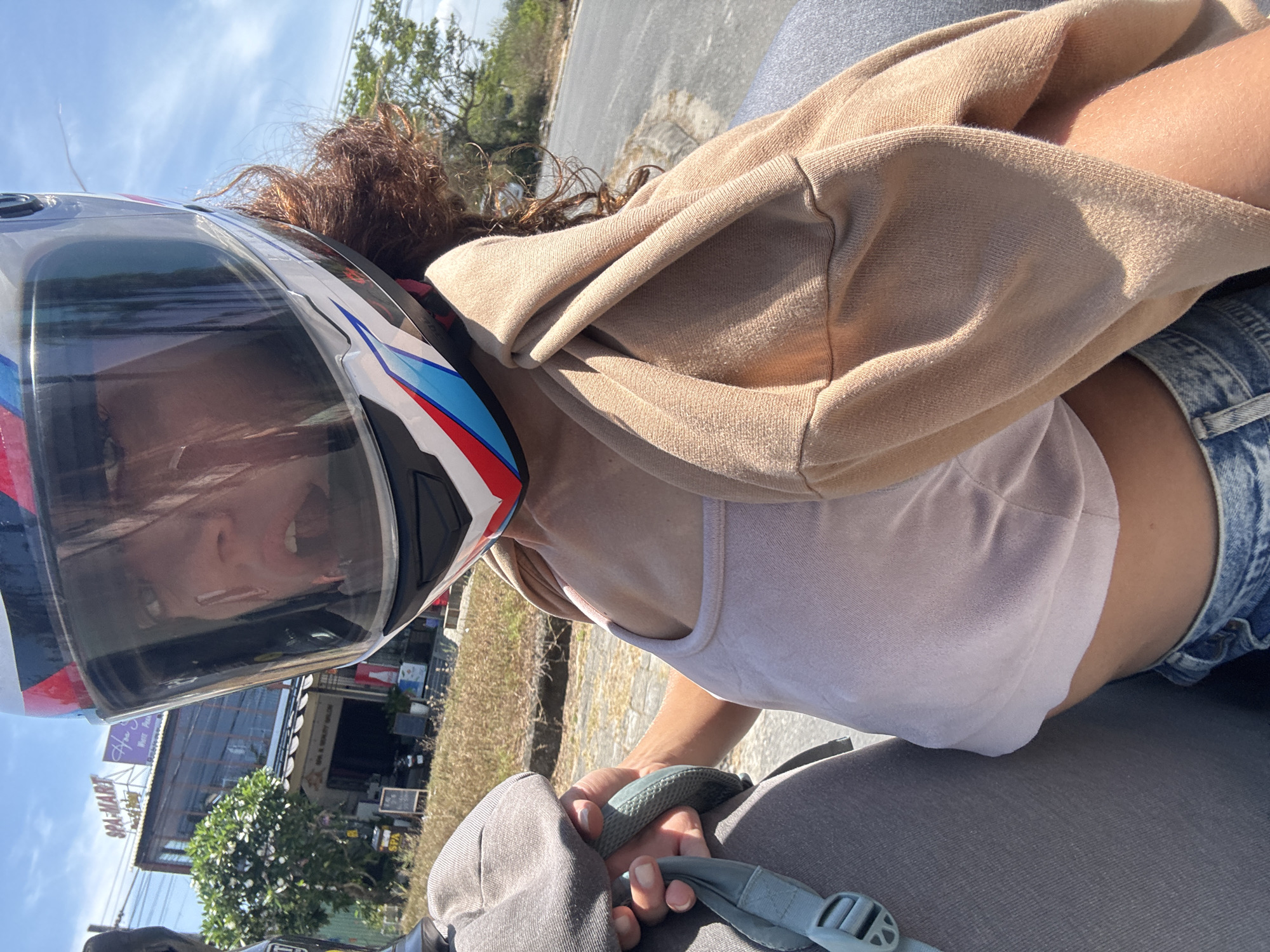 A woman wearing a motorcycle helmet is on the back of a motorbike, seemingly on the move, given the angle of the road and visible buildings. She is wearing a tan-colored hoodie over a white tank top and denim shorts. Her brown hair is visible under the helmet. She is holding onto a gray backpack with light blue straps. The background shows buildings, including one with a sign reading "SPA-MARKET" and "Apina Wave Pool & Beach Club Ware House." The location is identified as Đà Nẵng, Vietnam. The time of day appears to be daylight, with a bright sky and sunlight present.