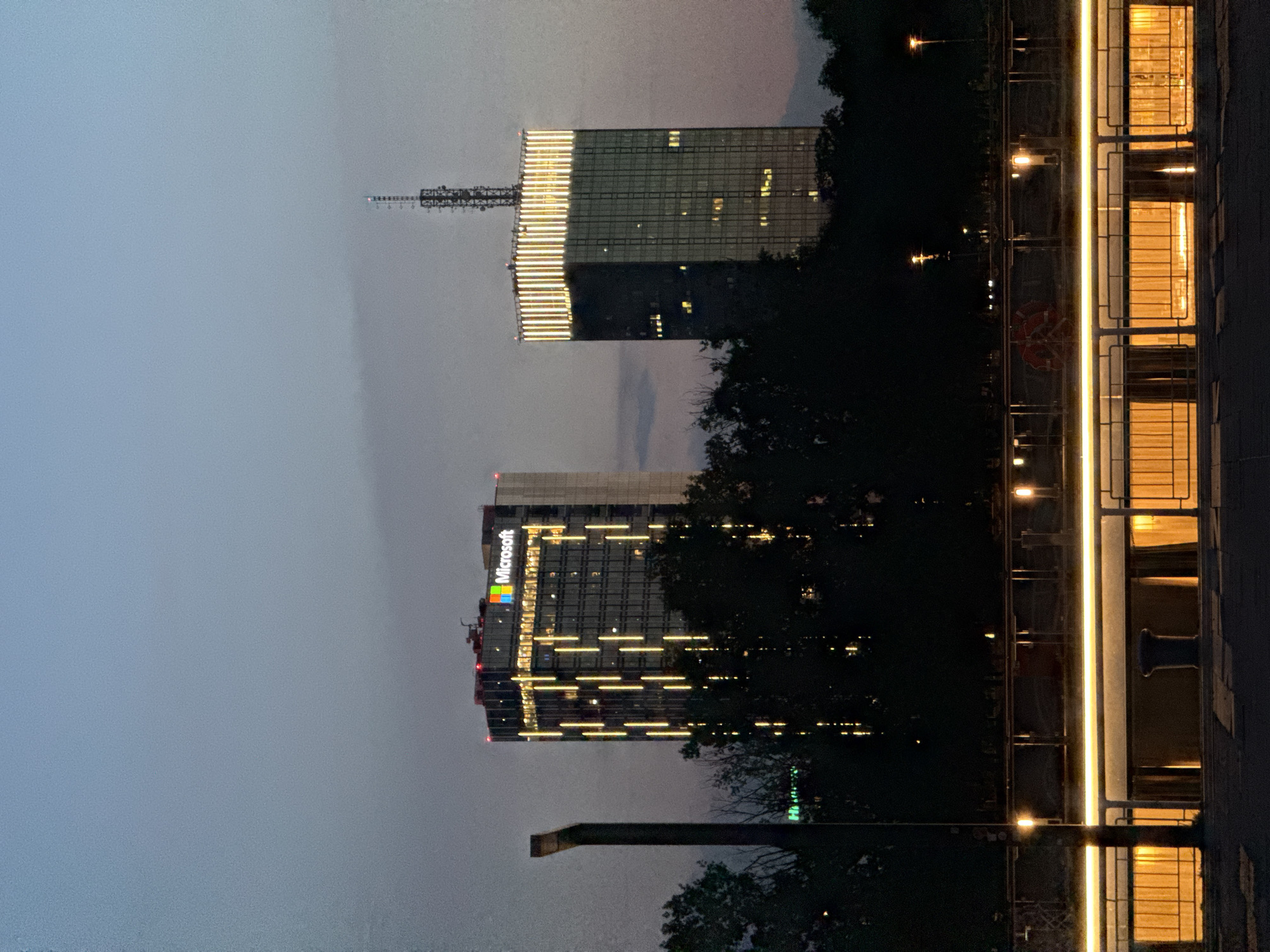 This image depicts the New Belgrade skyline at dusk, with a focus on the Western City Gate (also known as Genex Tower) and the nearby Microsoft building. The sky is a gradient of deep blue and purple, indicating the transition from day to night.
The Western City Gate, a prominent landmark in Belgrade, is on the right side of the frame. It is a tall, modern skyscraper characterized by its glass facade. The upper section of the tower is illuminated by a series of vertical yellow lights, creating a striking visual effect against the darkening sky. A communication antenna is visible at the very top of the tower.
To the left of the Genex Tower is the Microsoft building, another skyscraper. It features a glass facade with numerous illuminated windows that create a grid-like pattern. The building is adorned with vertical lines of warm yellow lights that descend from its upper floors, adding to the nighttime ambiance. The "Microsoft" logo, complete with its colorful quadrangle, is prominently displayed on the upper section of this building.
In the foreground, silhouetted trees obscure the lower portions of the buildings, adding depth to the scene. Below the trees, a bridge or elevated walkway is visible, lined with railings and illuminated by a continuous strip of warm, golden light. Further down, beneath the walkway, is a row of structures with open windows, each emitting a warm orange glow, suggesting interior lighting.
A dark, vertical structure, possibly a chimney or a piece of industrial architecture, stands to the left of the Microsoft building, partially obscuring it. The overall impression is one of a modern urban landscape at twilight, with artificial lights contrasting against the natural fading light of the sky. There are no people or animals visible in the frame. The scene is static and captures a moment of urban serenity as night descends.
