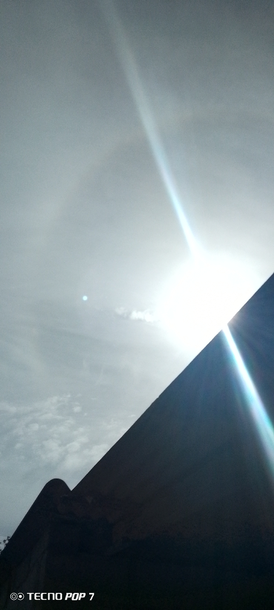 The image is taken outdoors during daylight, featuring a bright sun obscured partially by what appears to be a building or structure. The sky is overcast with visible clouds. The lower portion of the image shows the silhouette of a dark structure that could be a building or a wall. The bottom left corner has "TECNOP POP 7" printed, suggesting the type of device used to capture the picture. Without further context, I am unable to provide details about the city or specific location in Altagracia, Venezuela.