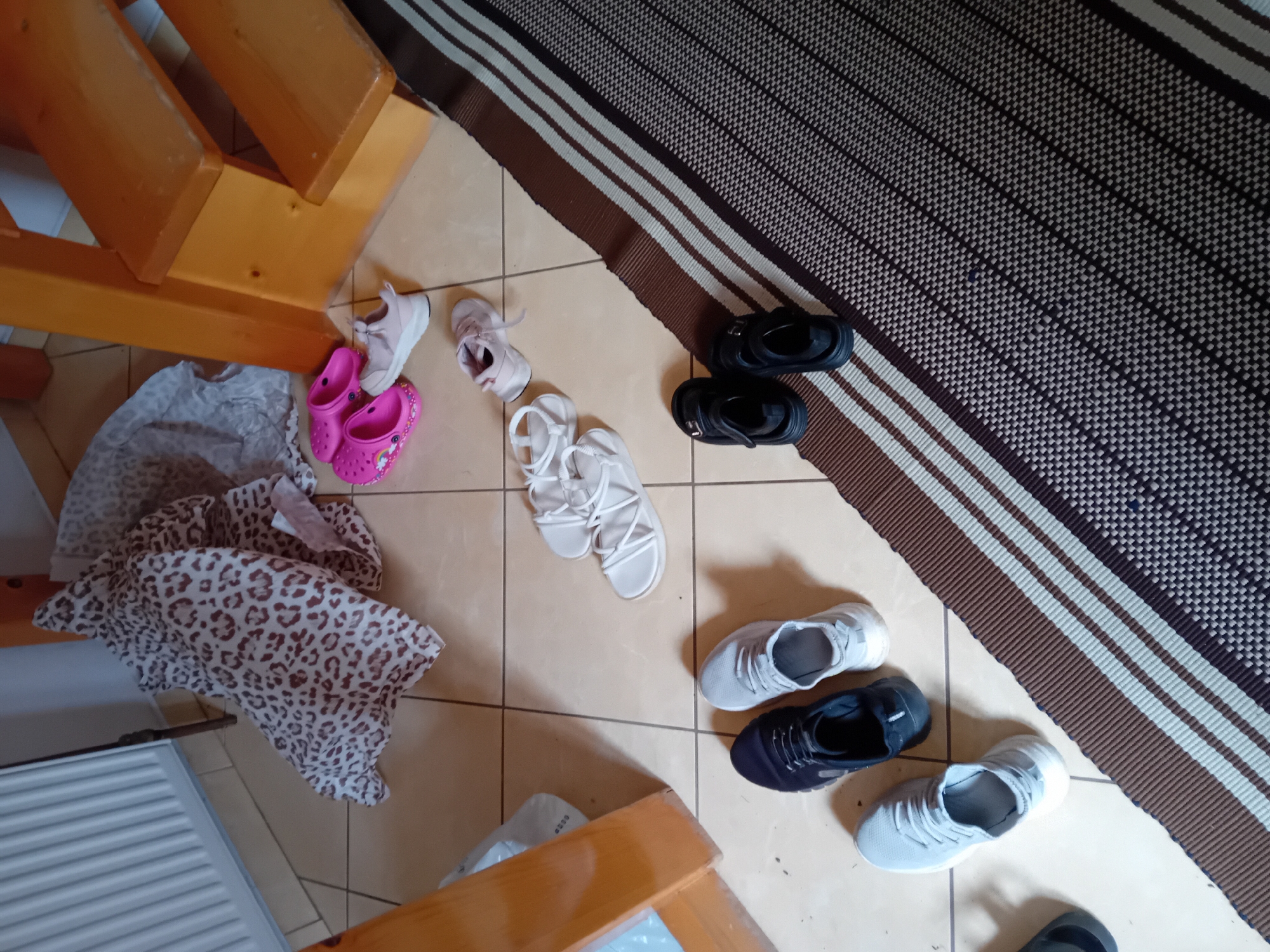 The image depicts a cluttered floor area within a building, possibly a home, in Zuberec, Slovakia. The floor is tiled with large, light-colored square tiles. In the background, a wooden structure, likely stairs or furniture, is visible. To the left, a white radiator is present. Various footwear items are scattered across the floor, including several pairs of sneakers in shades of grey and dark blue, white sandals, pink Crocs, and black sandals. A patterned fabric with a leopard-like print is draped over some of the items. A striped rug runs diagonally across the lower right portion of the image. The scene appears to be an indoor domestic environment where shoes have been removed and left in disarray.