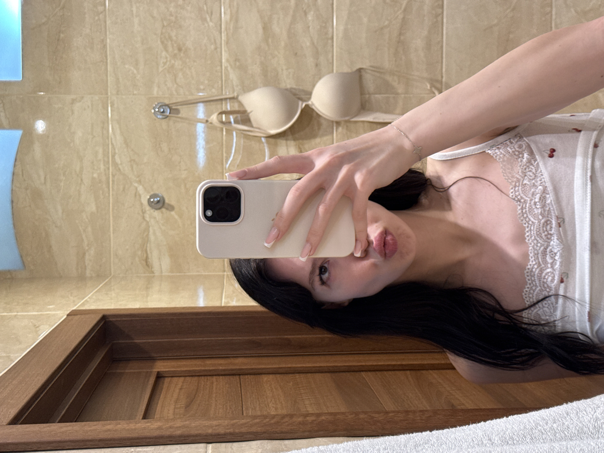 A young woman with dark hair is taking a selfie in a bathroom. She is wearing a white camisole with lace trim and a cherry print. She is puckering her lips and looking into the camera. In the background, there is a wooden door, tiled walls, and a bra hanging on hooks. The scene is consistent with a residential bathroom in Khimki, Russia.