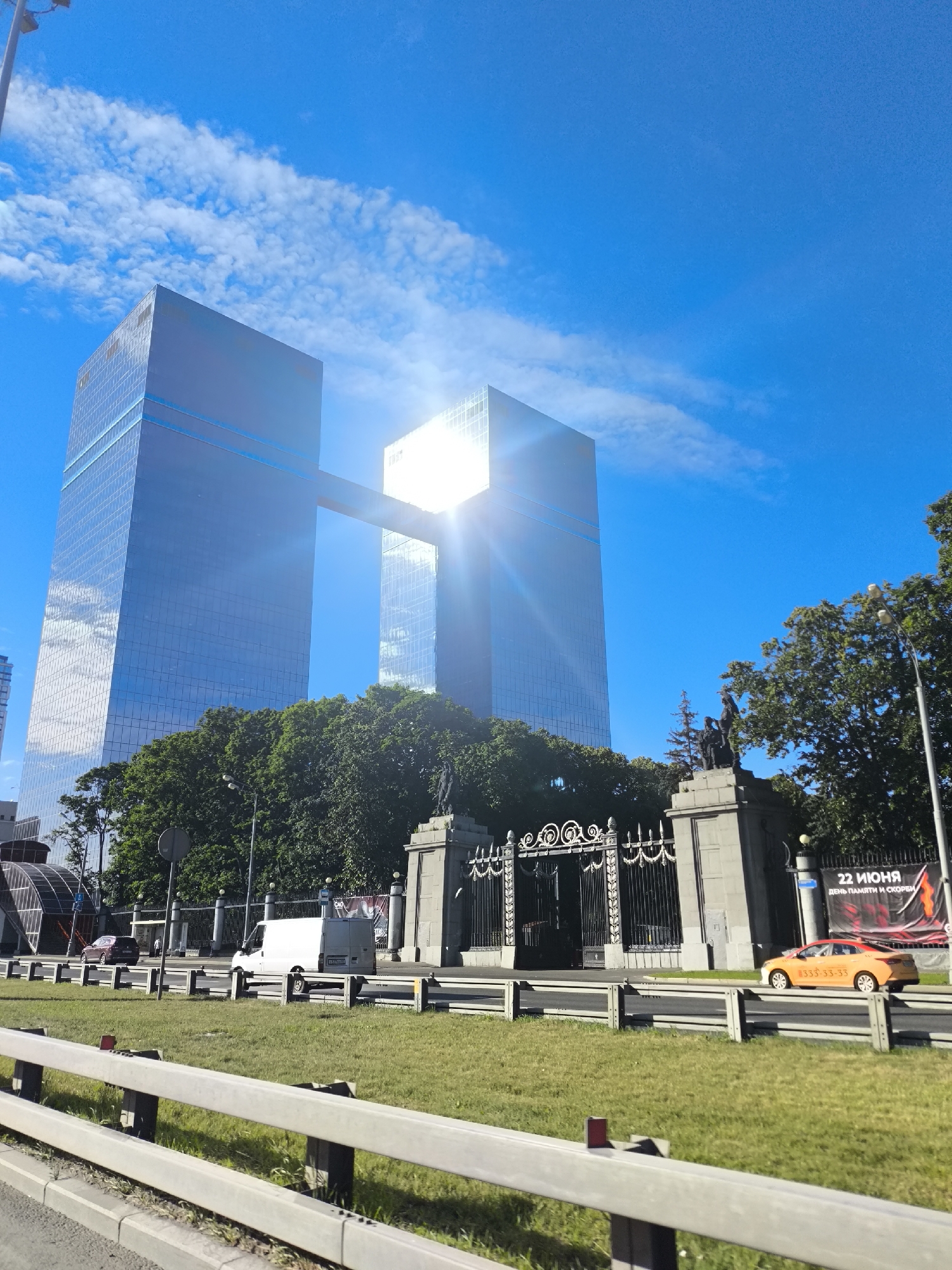 The image shows a sunny day in a city, with two tall glass skyscrapers connected by a skybridge. The skyscrapers are modern and reflect the bright blue sky and clouds. In front of the skyscrapers, there is a large, ornate gate with statues on top of it, surrounded by lush green trees. A road with a white van and an orange taxi is visible in the foreground, along with a grassy median and a white barrier. A banner with Russian text and graphics is hanging on the right side of the gate. The overall scene depicts a busy urban environment with impressive architecture and clear weather.