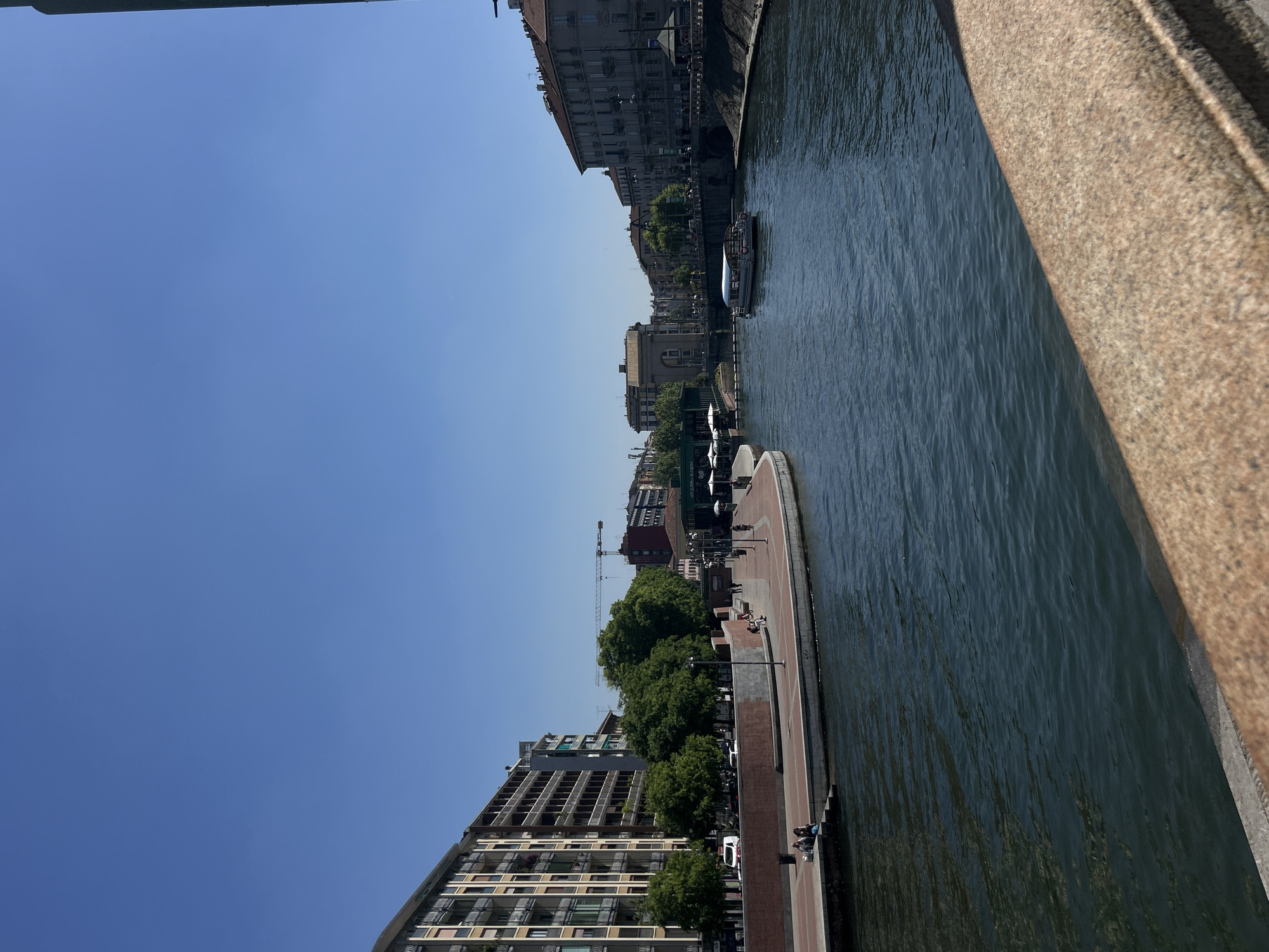 The image depicts a canal in Milan, Italy, likely the Naviglio Grande, viewed from a high vantage point such as a bridge. The water is visible, with subtle ripples suggesting movement. Buildings line both sides of the canal, varying in height and architectural style, ranging from older structures to modern apartment blocks. Trees provide greenery along the canal banks. A paved walkway runs along the canal. People are walking and congregating. A tour boat is moving on the water. The sky is clear and blue. Part of the bridge's structure is visible on the right side of the frame.