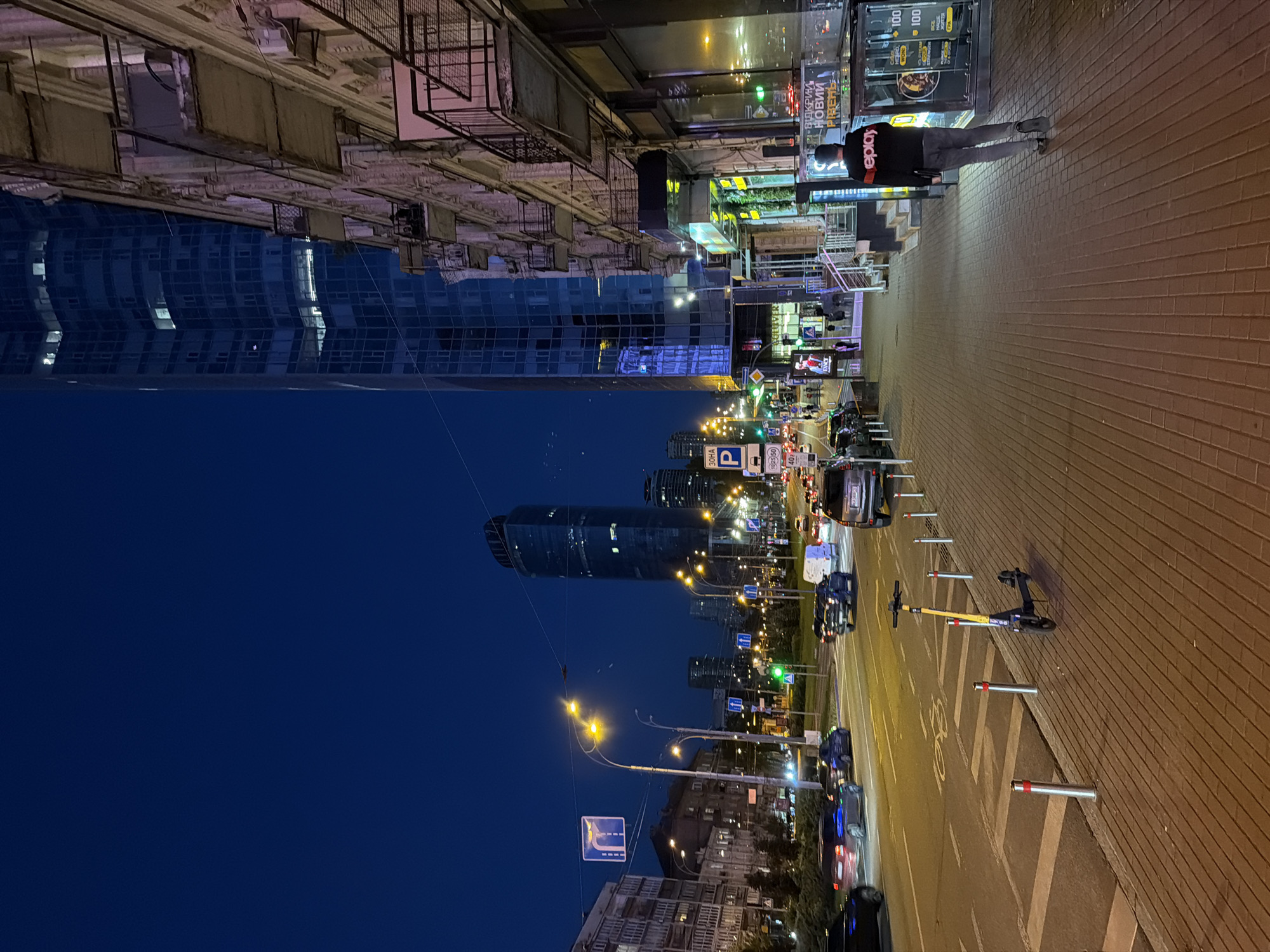 The image captures a nighttime street scene in Kyiv, Ukraine, likely in the Podil district given the architectural style and the presence of modern skyscrapers alongside older buildings. A tall, modern glass skyscraper dominates the right side of the frame, its illuminated windows reflecting the dark blue twilight sky. Adjacent to it is an older, ornate building with balconies, showing signs of age. A wide road with multiple lanes is visible on the left, with cars in motion and traffic lights indicating normal flow. Streetlights and illuminated signs cast a warm glow on the scene. A person, possibly a young man, walks away from the camera down the brick-paved sidewalk on the right. He is wearing a dark sweatshirt with a white logo on the back and dark pants. A yellow electric scooter is parked on the sidewalk near the street. The overall atmosphere is that of a bustling urban environment at night, with a mix of old and new architecture.