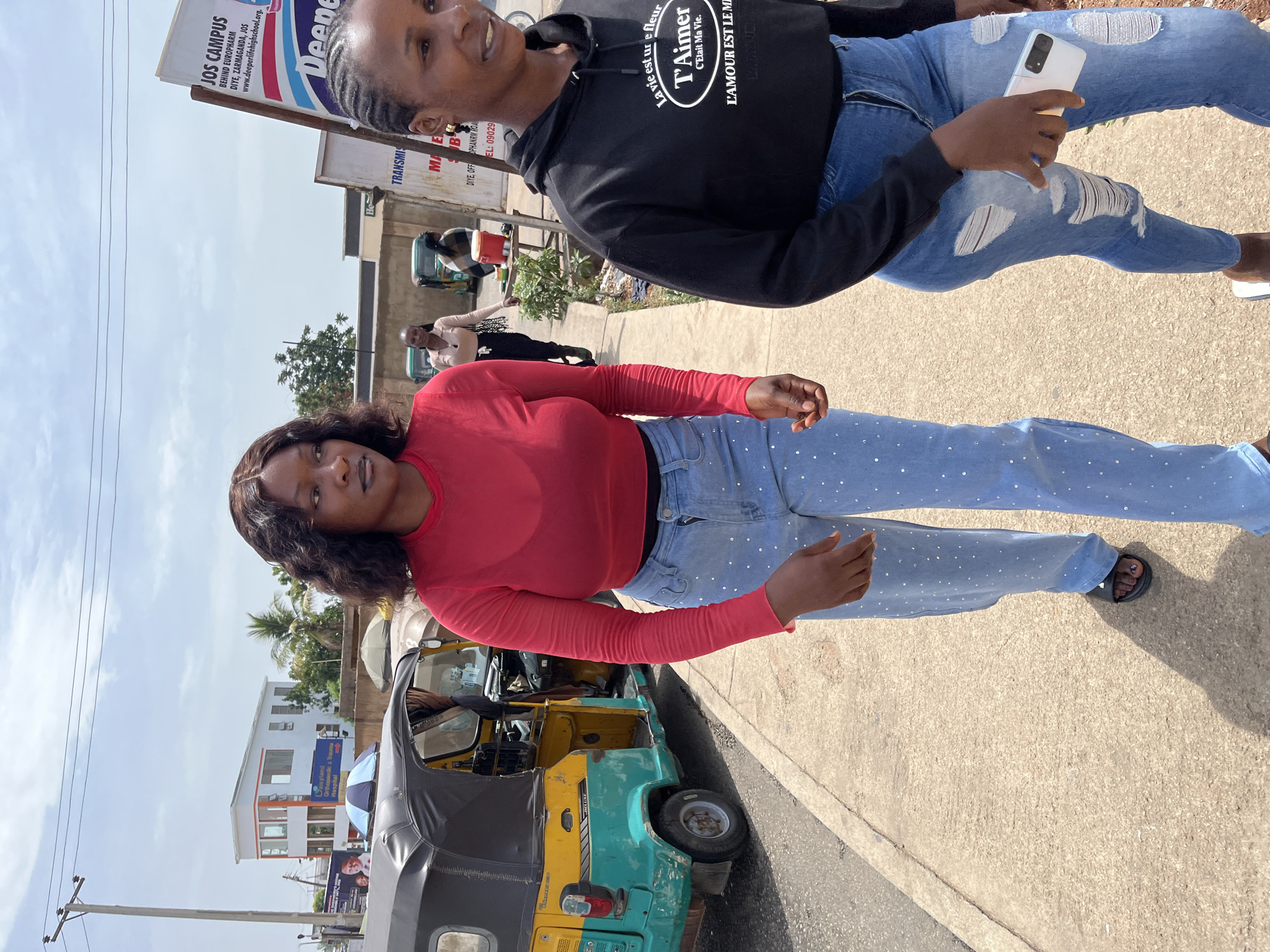 Two women are captured walking on a sunlit street in Jos, Nigeria. The image is taken from a slightly low angle, showing a bright, clear sky above.
In the foreground, a woman with dark, wavy hair wears a red long-sleeved top and light blue jeans adorned with small white embellishments. Her left foot, wearing a dark sandal, is visible. She looks towards the camera with a neutral expression.
Behind her, another woman with dark braided hair smiles towards the camera. She is dressed in a black hoodie featuring white text including "La vie est une fleur," "T'Aimer C'Etait Ma Vie," and "L'AMOUR EST LE MIEUX." She wears blue ripped jeans and holds a white smartphone in her right hand.
To the left of the foreground woman is a yellow and teal/green tricycle (keke napep) parked on the roadside. The background reveals various street signs and buildings. Notable visible text from signs includes "JOS CAMPUS," "BEHIND EUROPHARM," "DIYE, ZARMAGANDA, JOS," and "www.deeperlifehighschool.org." Another sign indicates "Daisyland Orthopaedic Hospital," and further text like "TRANSMIS MALE B" and a phone number "EL: 09029" are also partially visible.
The scene depicts a casual daytime moment, with a few other people partially visible in the background, suggesting a moderately busy street. The overall atmosphere is relaxed and sunny.
