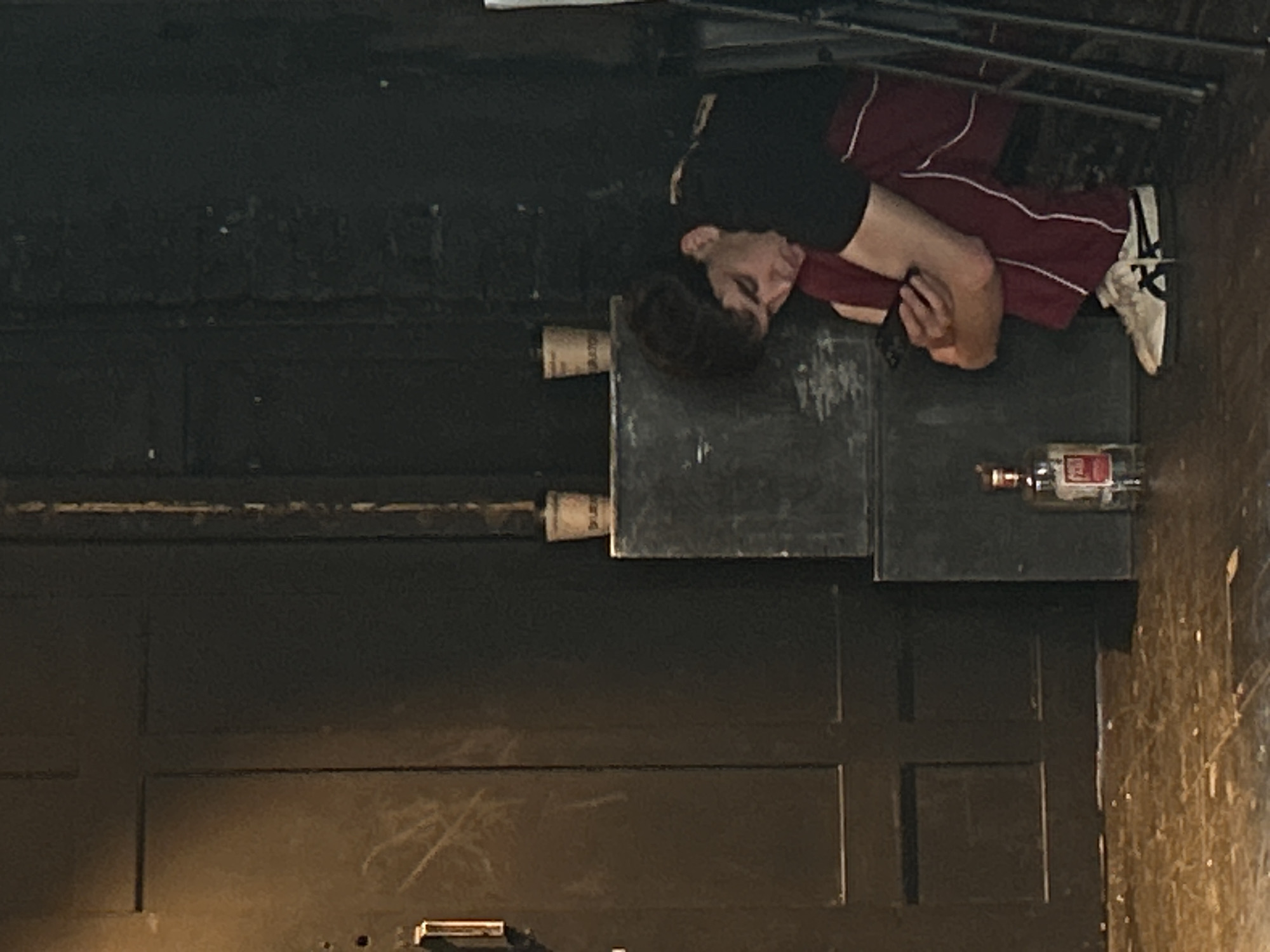 A man with short dark hair, wearing a black short-sleeved shirt and maroon pants with white stripes, lies prone with his head resting on a set of three stacked dark gray blocks on a dark wooden floor. Next to the blocks, there is a clear glass bottle containing liquid with a red label. Nearby there are two rolled paper tubes taped to the side of another dark gray block. A metal folding chair is visible in the top right corner of the frame. The setting appears to be an indoor space with dark paneled walls. The location is listed as Unknown, Unknown.