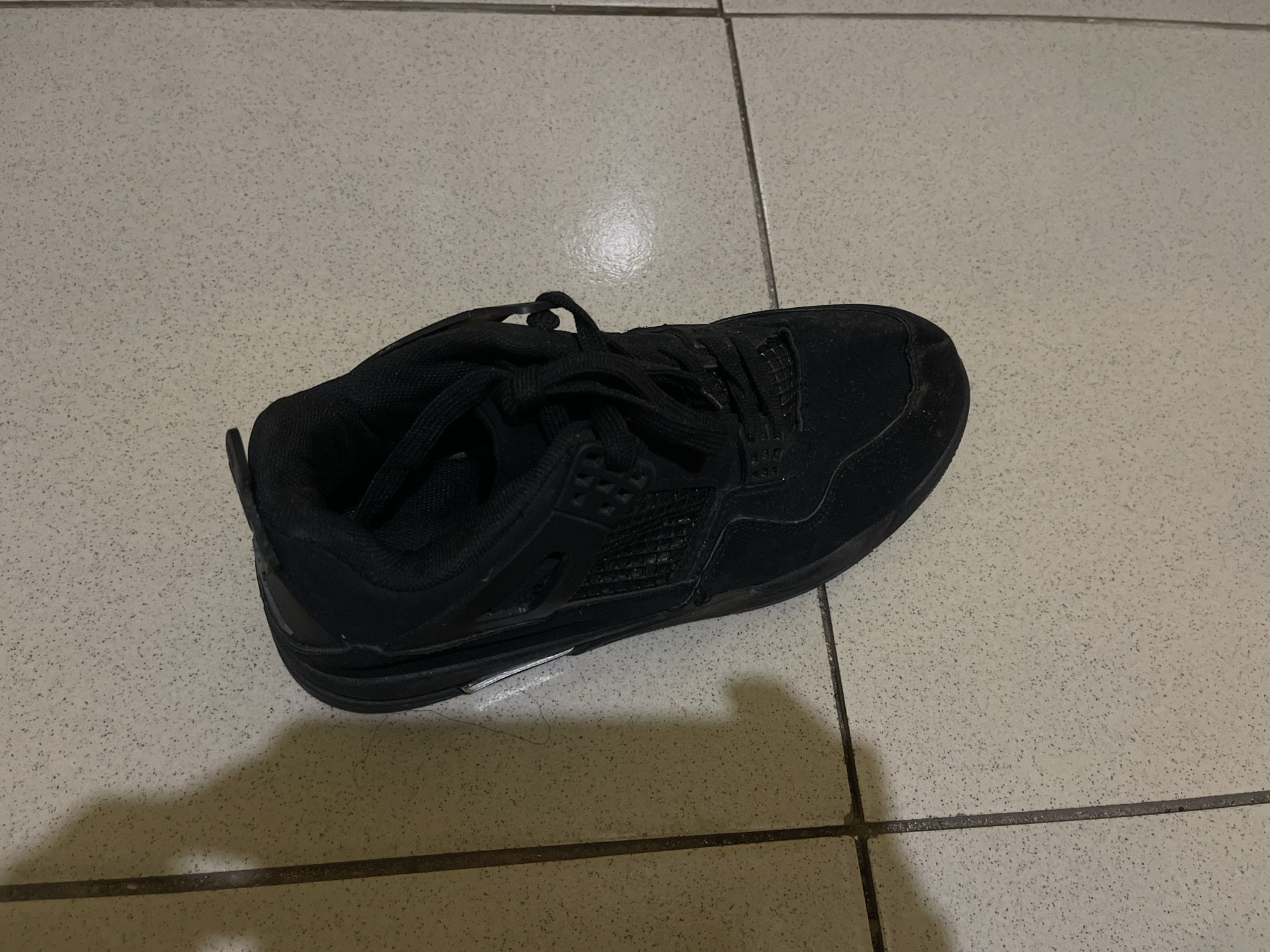 A single, black athletic shoe sits on a tile floor. The shoe is laced and appears to be a low-top model. The tiles are light in color with small, dark speckles. The location is unknown, and no people are visible. The presence of the shoe on the floor suggests it has been placed or dropped there.